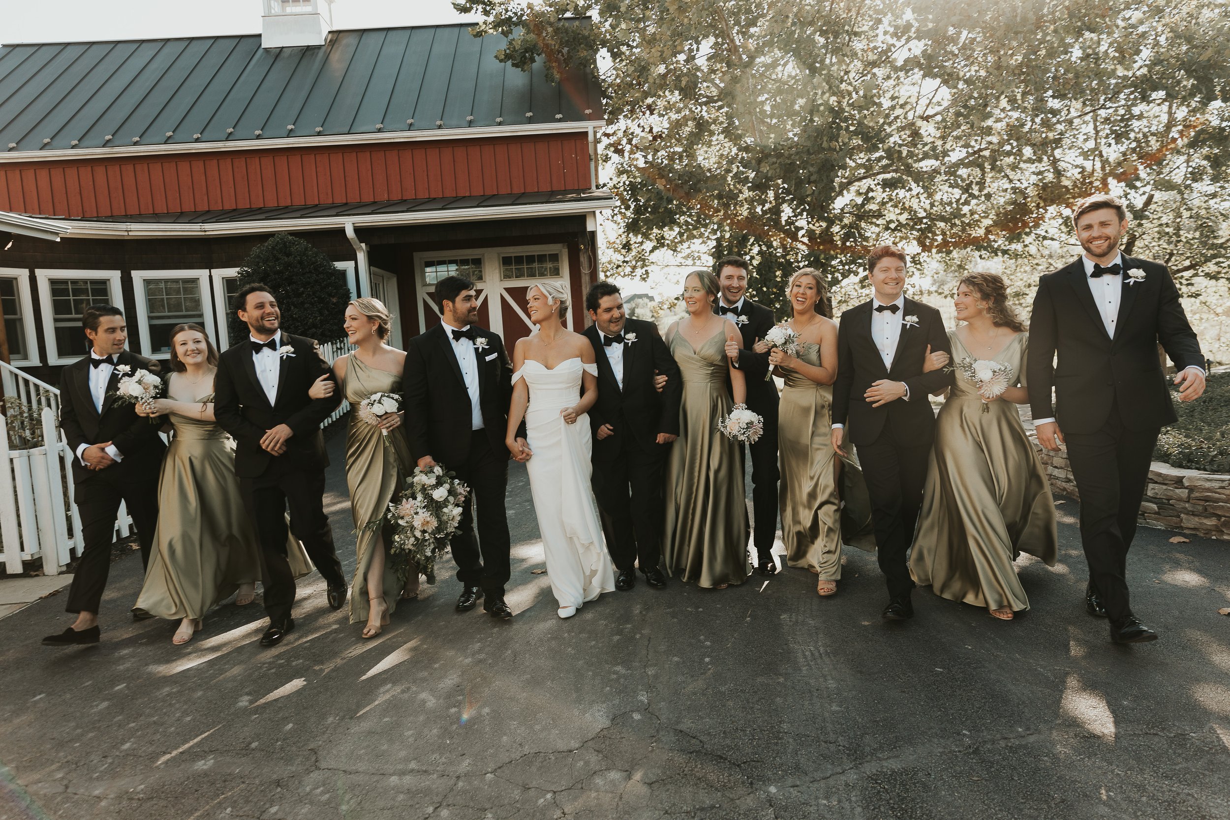 A group of people dressed in formal attire walking outside in front of a house with a green roof and a tree, celebrating a wedding.