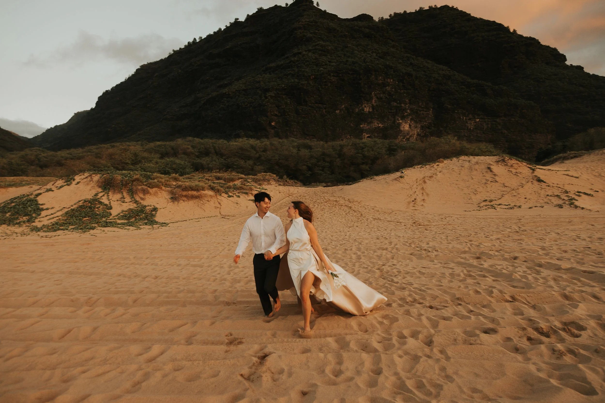 A destination wedding couple in wedding attire walking and holding hands on a sandy beach at sunset, with a large mountain in the background.