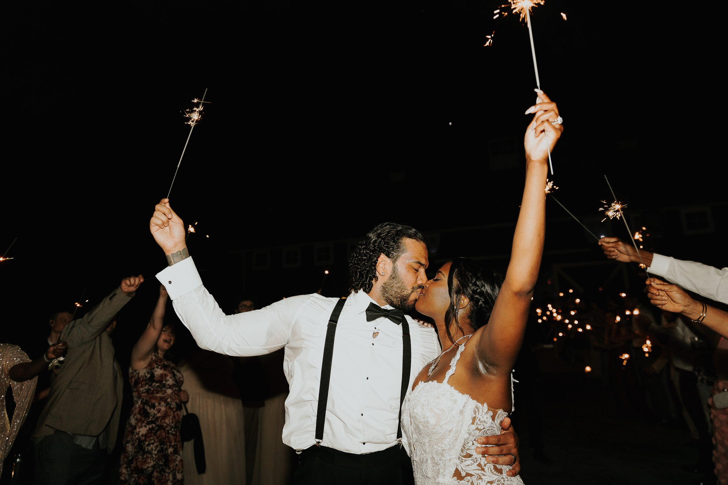 A couple is kissing during a celebration at night, holding sparklers in the air with people dancing and celebrating around them.
