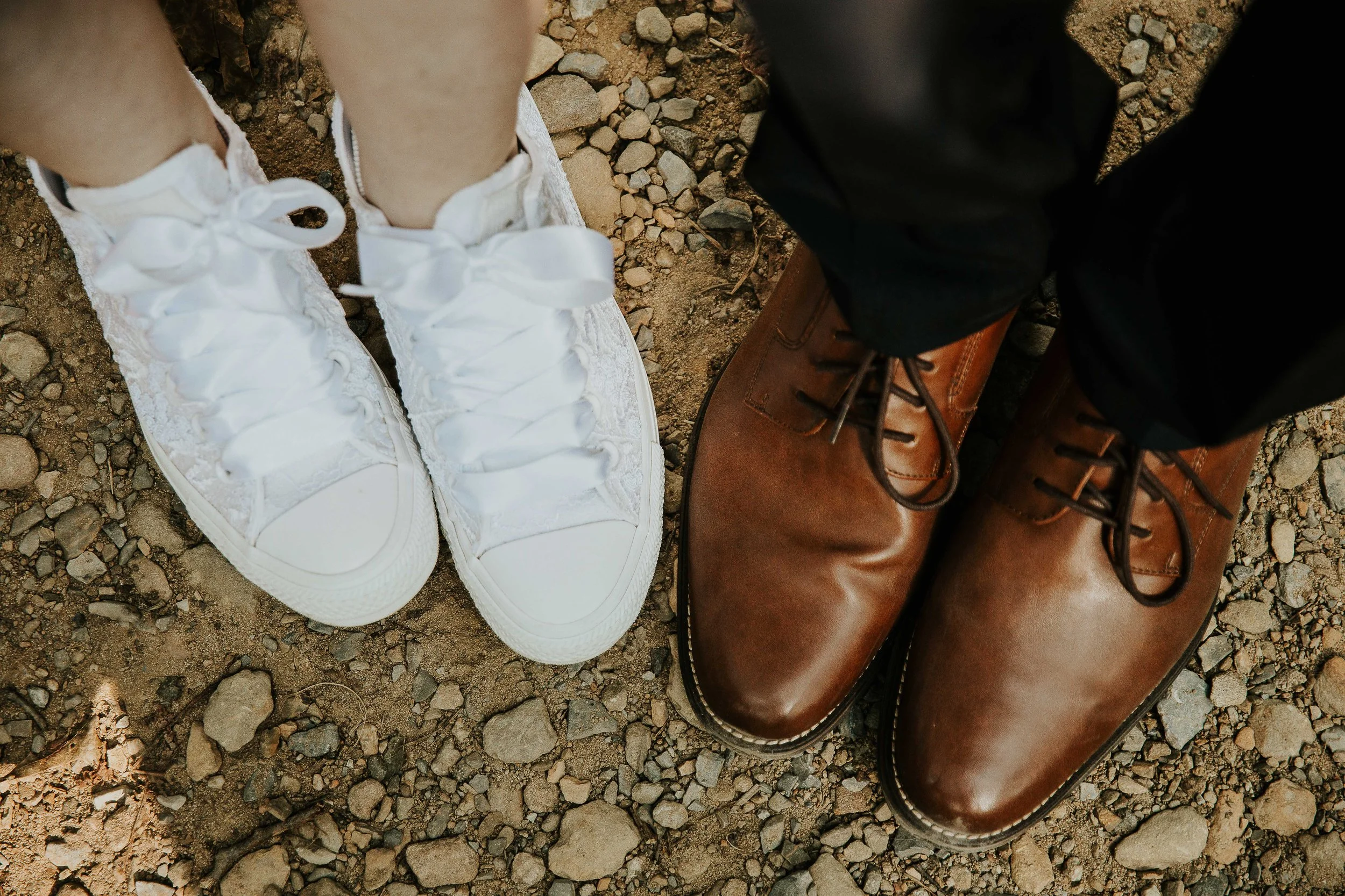 Close-up of two pairs of shoes on gravel ground. On the left, a person is wearing white lace-up sneakers with large satin bows. On the right, another person is wearing brown leather boots with black laces, and black pants.