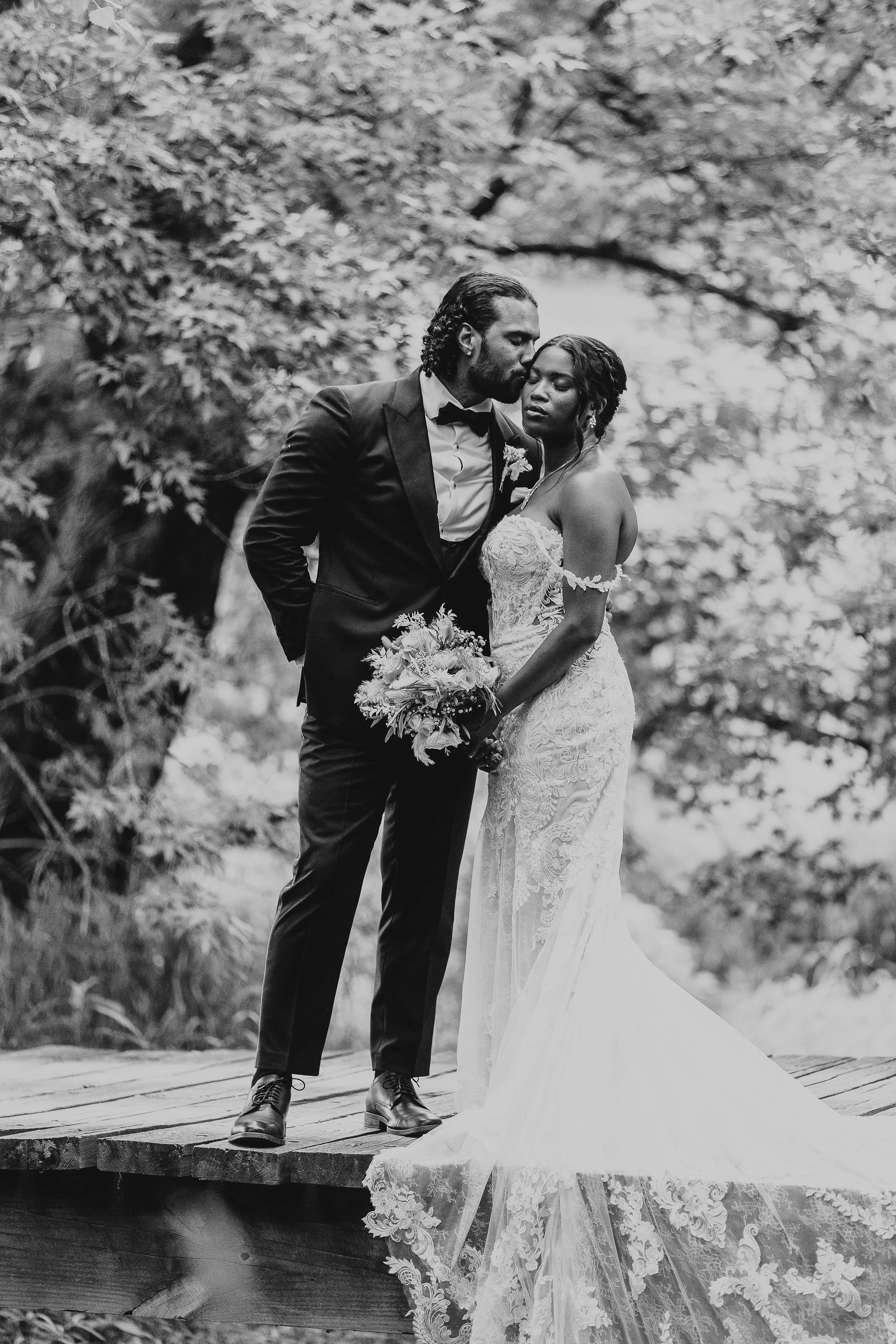 A black and white photo of a bride and groom standing on a wooden platform outdoors, with a forest and water in the background, sharing an intimate moment.