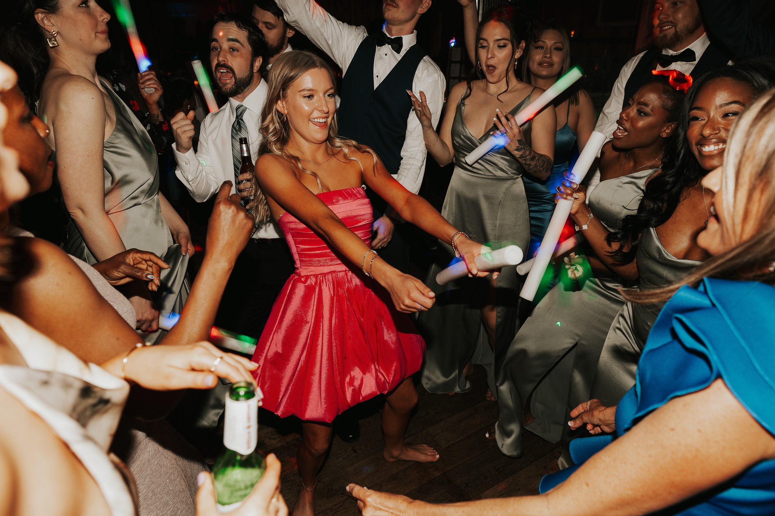 Group of people dancing and enjoying at a party, with some holding glow sticks and beer bottles.