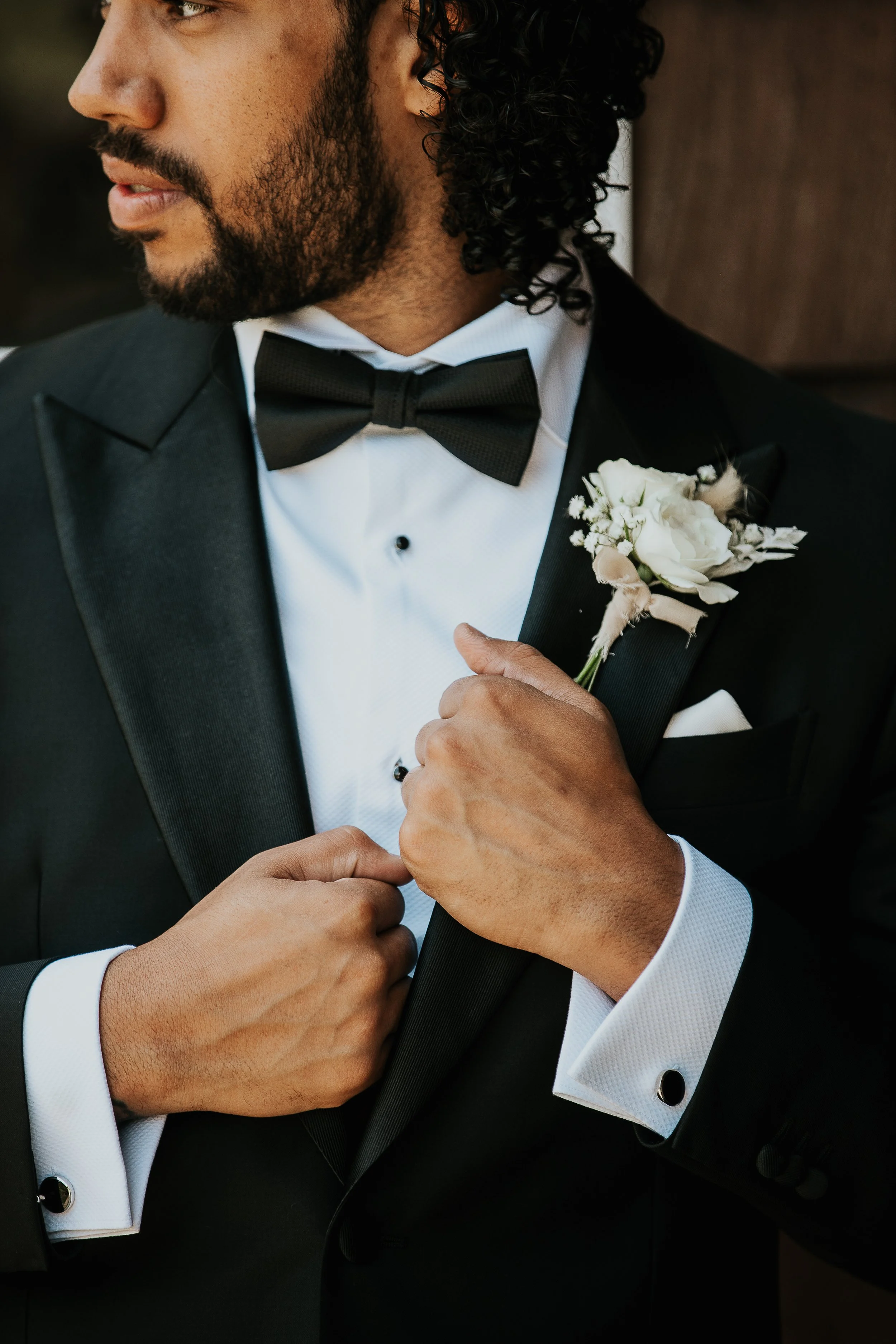 A man in a tuxedo, adjusting his bow tie and holding a boutonniere of white flowers on his lapel.
