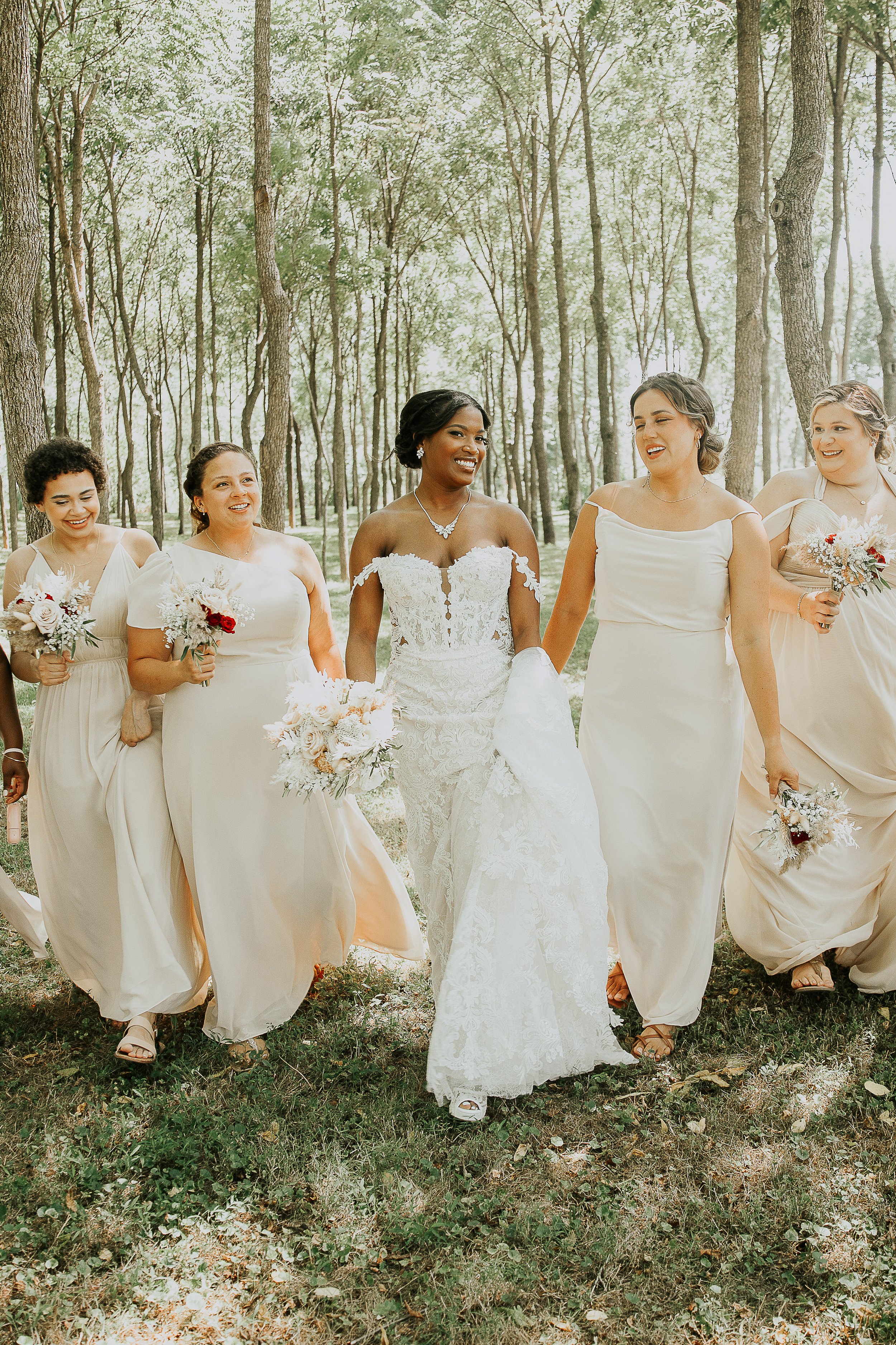 A diverse group of women in wedding dresses walking through a wooded outdoor area, smiling and holding bouquets.