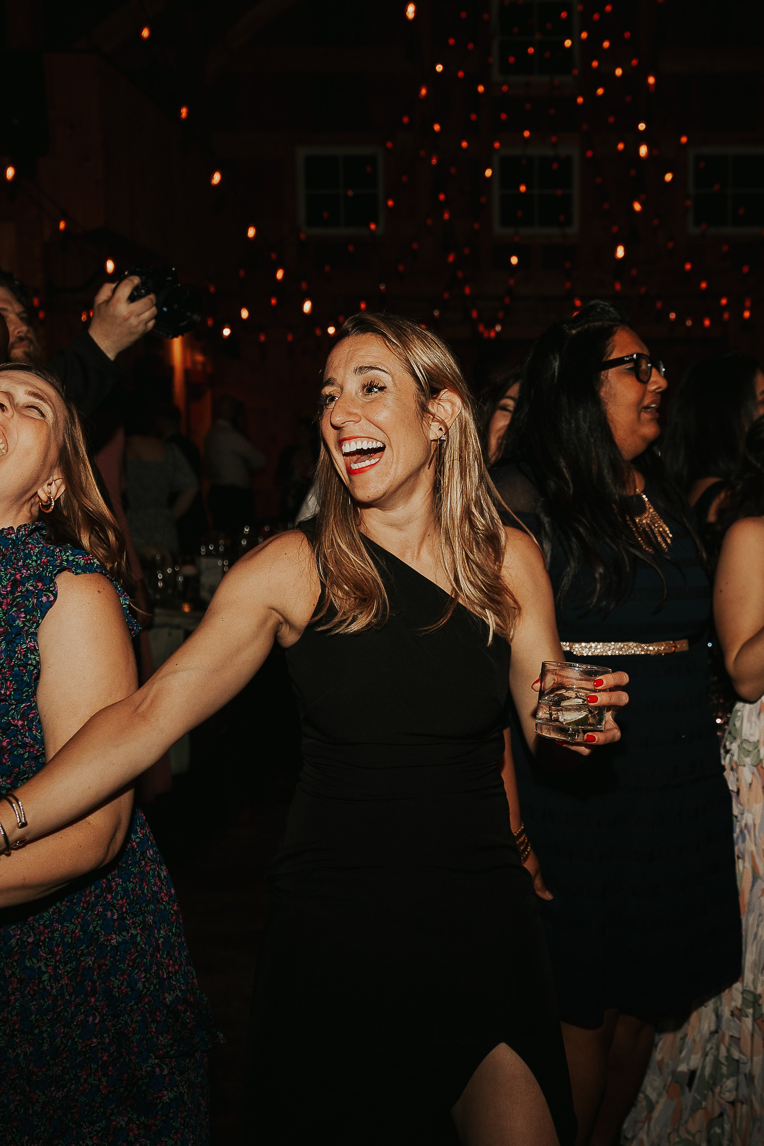 A group of women at a social gathering or party in a dimly lit venue, enjoying themselves and smiling. One woman in the center is holding a drink and appears to be laughing.