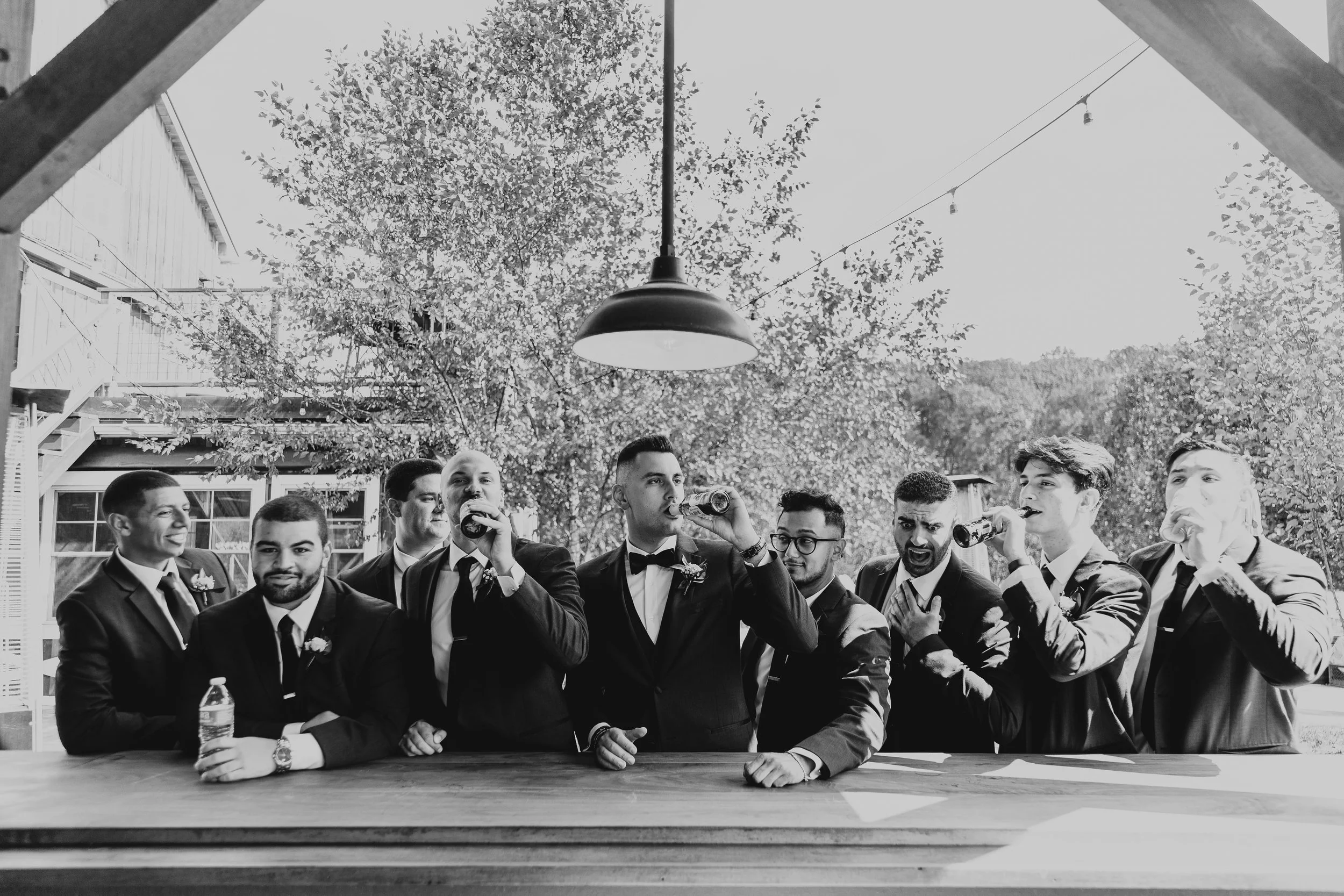 A group of men in formal suits and ties, some drinking beer, standing outdoors under a large tree with a house and string lights in the background, captured in black and white.
