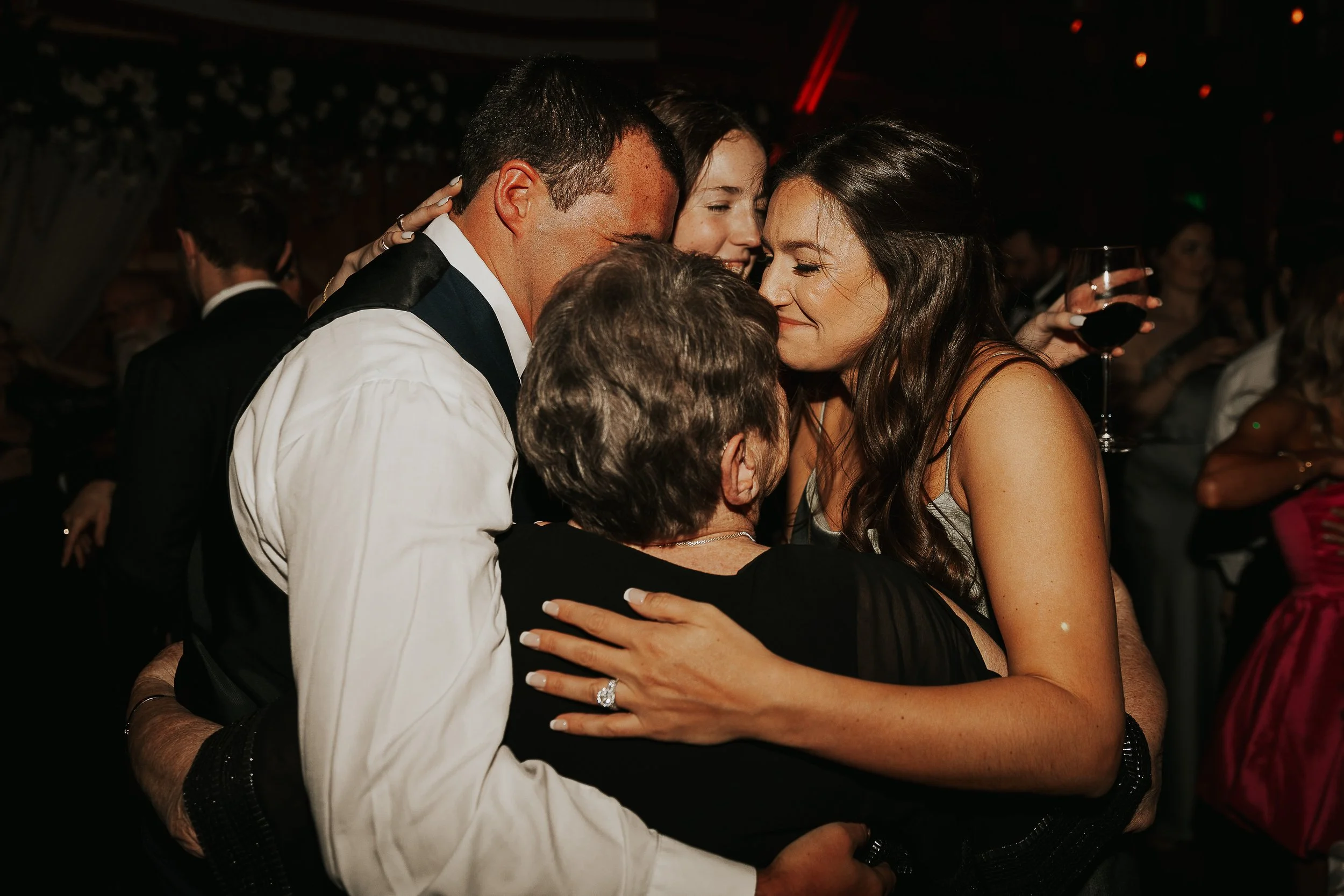 Four people hugging at a party, with a woman holding a glass of red wine in the background.
