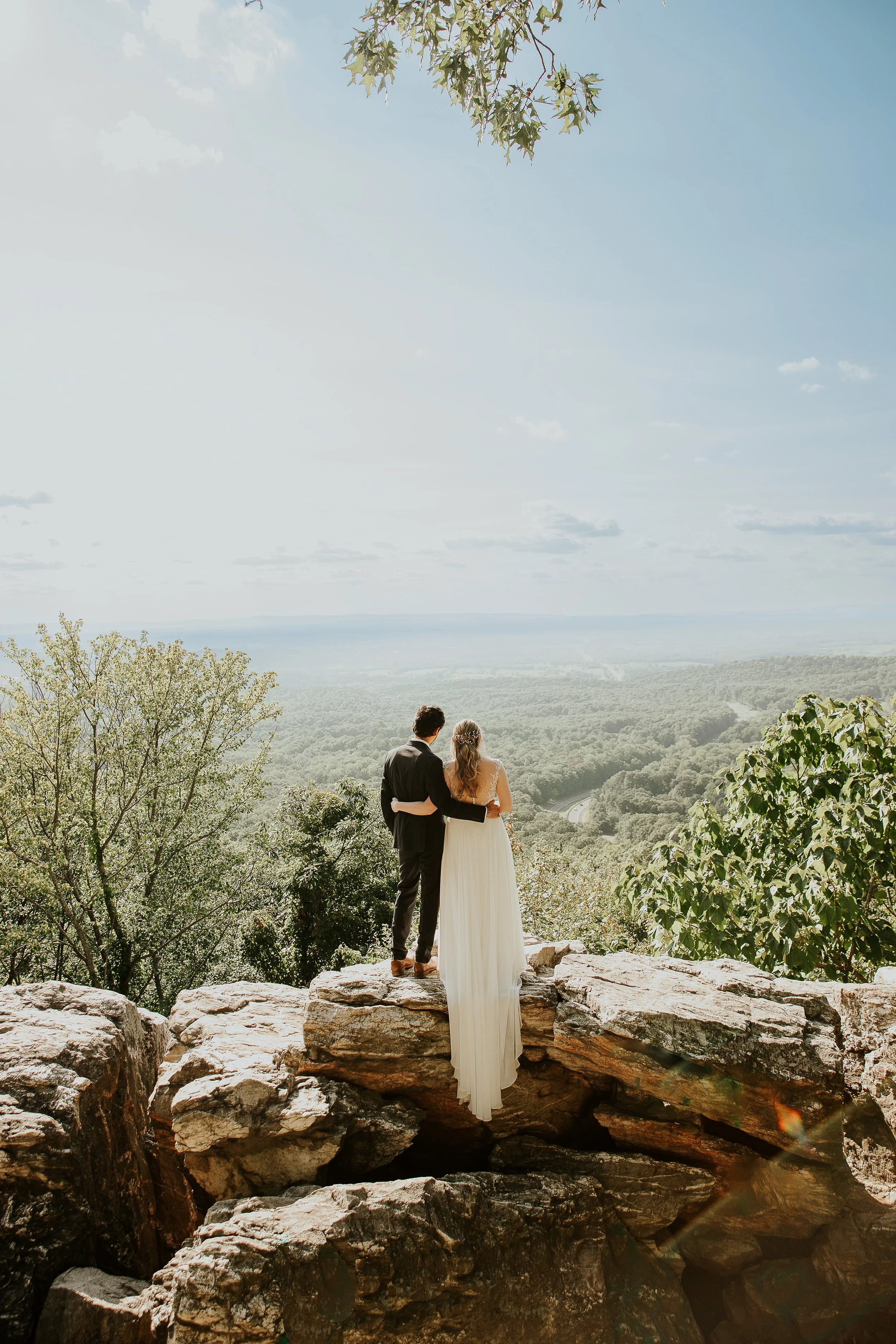 An elopement couple in wedding attire stands on a rocky ledge overlooking a vast landscape of trees, fields, and a distant river or road under a partly cloudy sky.