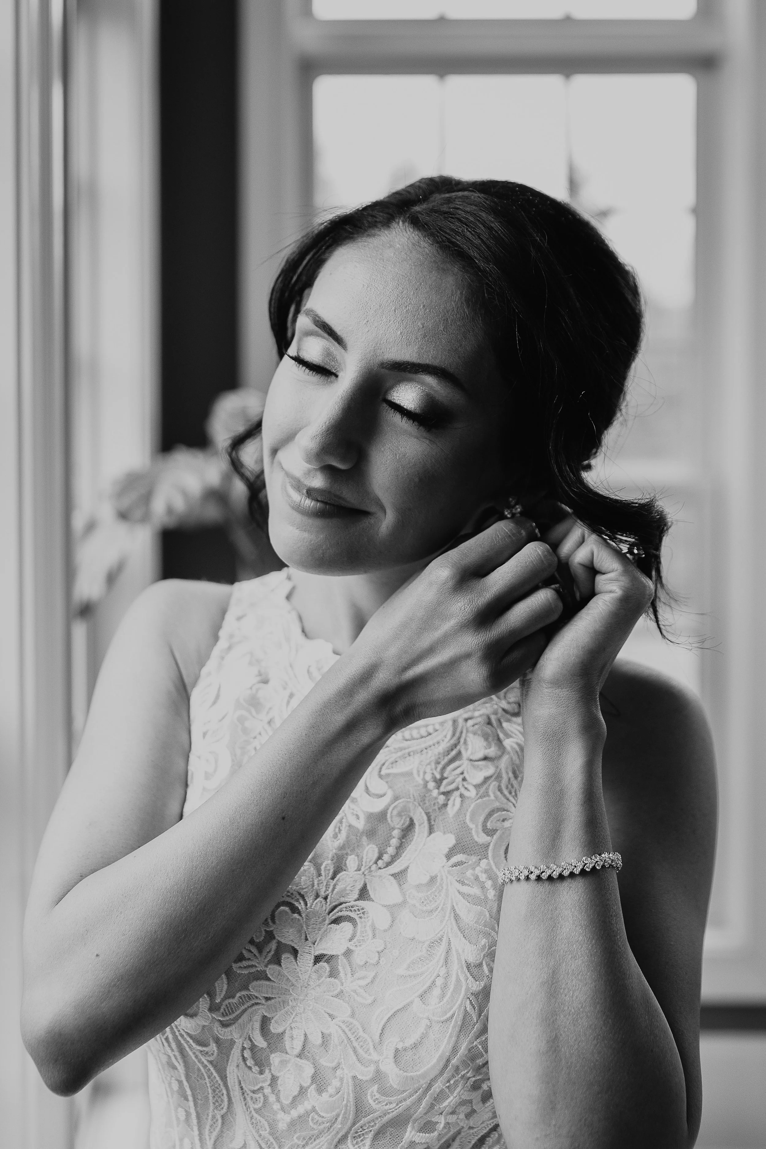 A black and white photo of a woman with dark, wavy hair, and closed eyes, adjusting an earring. She is wearing a lace dress and a bracelet, standing in front of a window.
