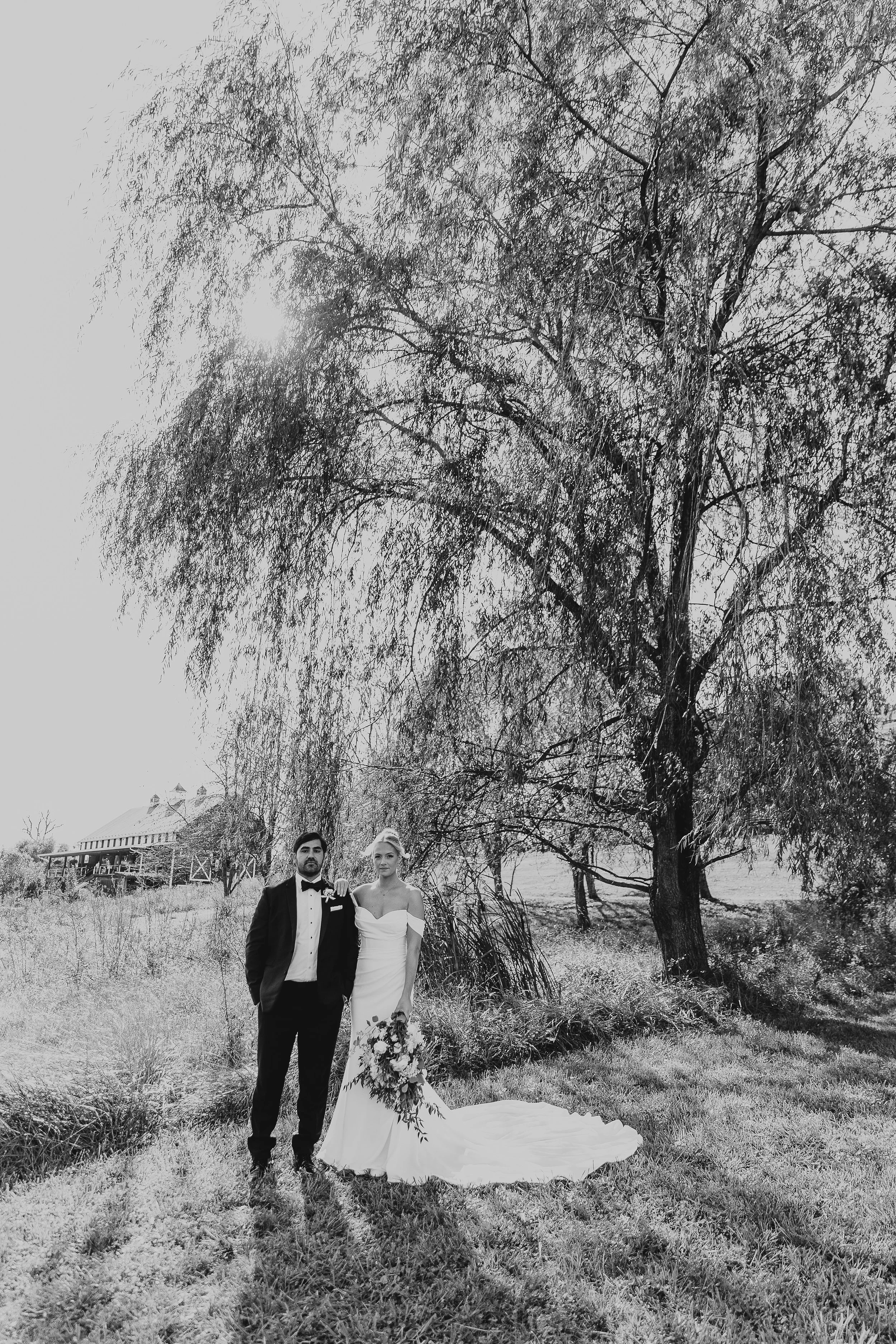 A bride and groom standing outdoors on grass with large trees in the background. The groom wears a tuxedo, and the bride wears a wedding dress and holds a bouquet. There is a building with a porch in the distance.