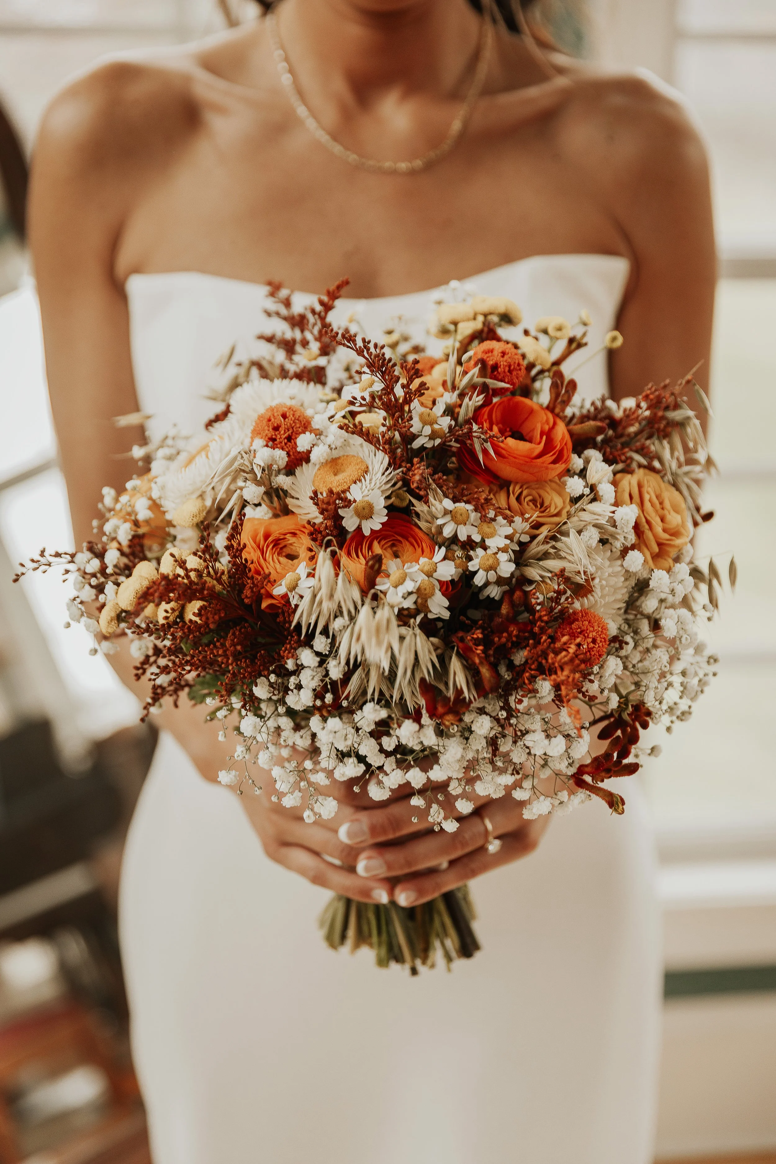A bride holding a bouquet of orange, white, and cream-colored flowers, wearing a strapless white wedding dress and a pearl necklace.