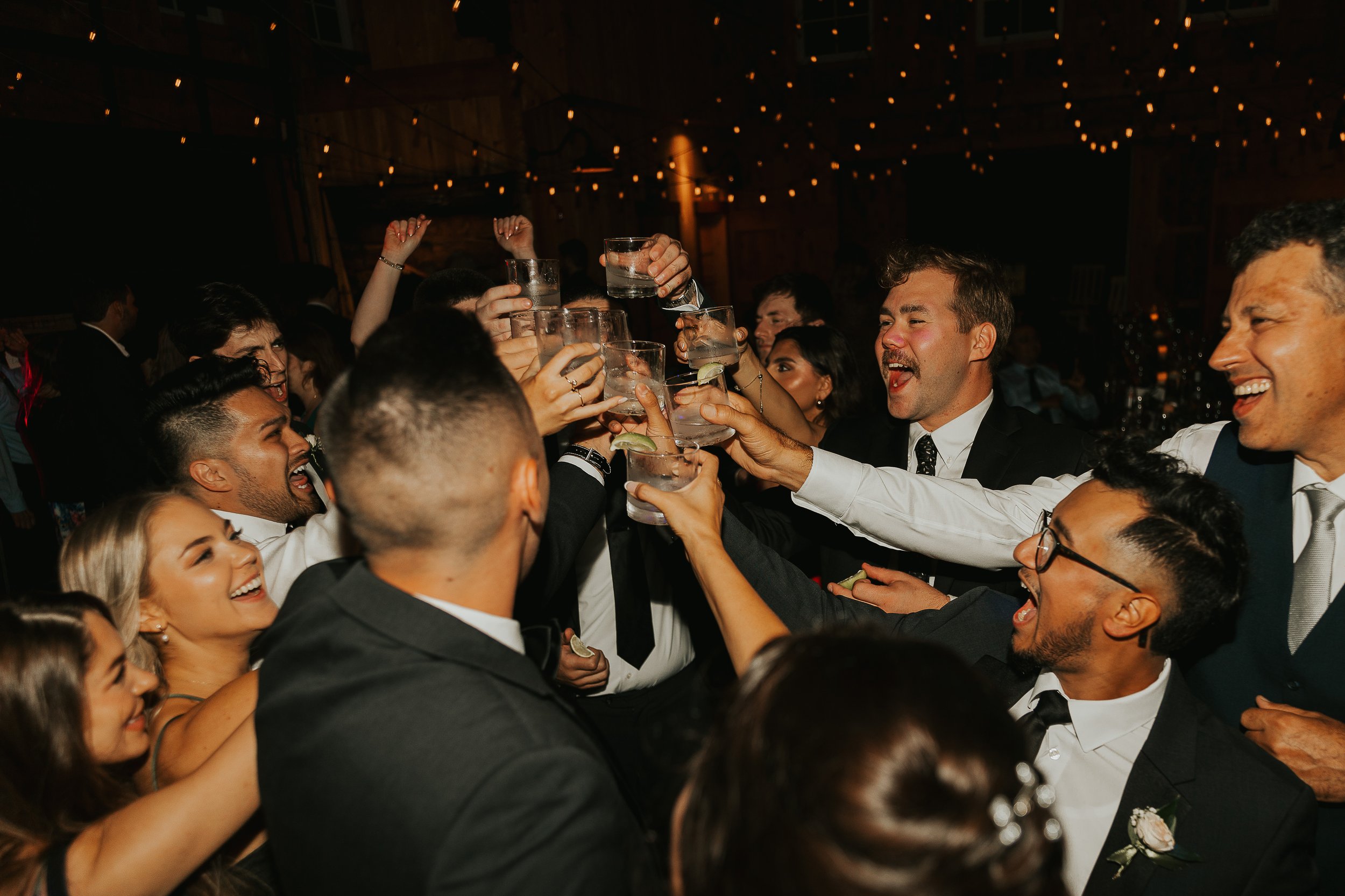 A group of people dressed in formal attire celebrating at a party, raising glasses for a toast in a dimly lit venue with string lights overhead.