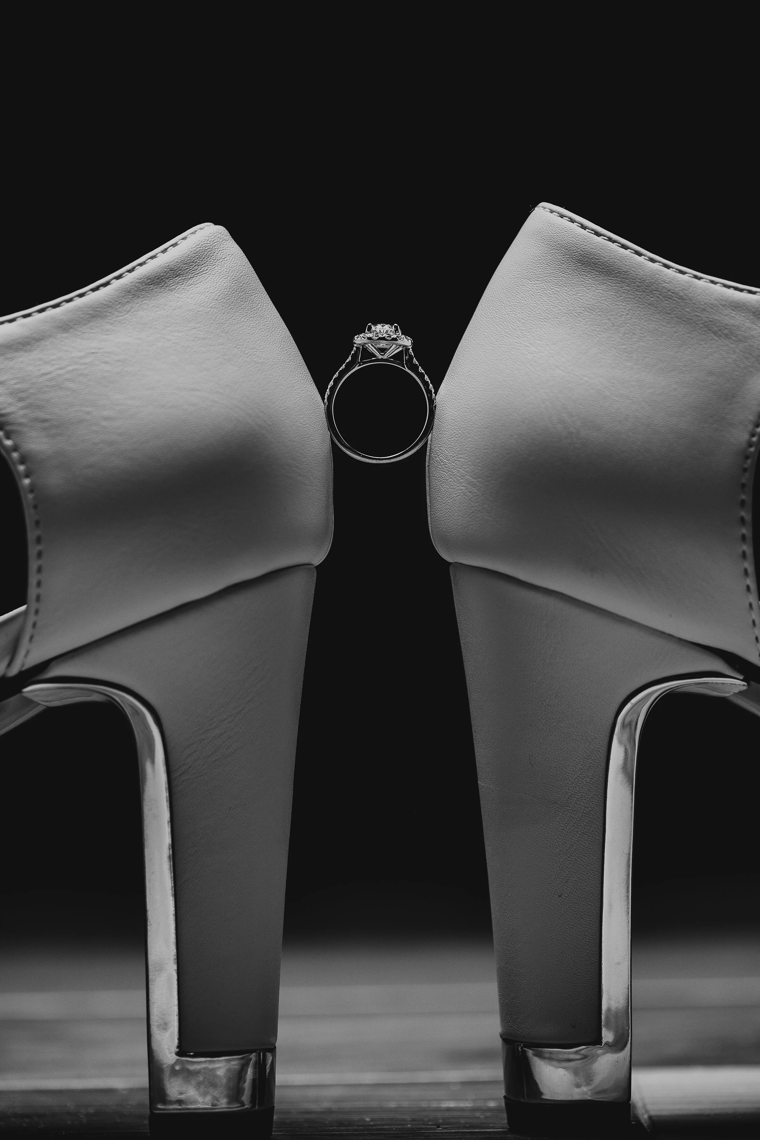 Close-up of a pair of high heeled shoes with a ring placed centered between them against a dark background.