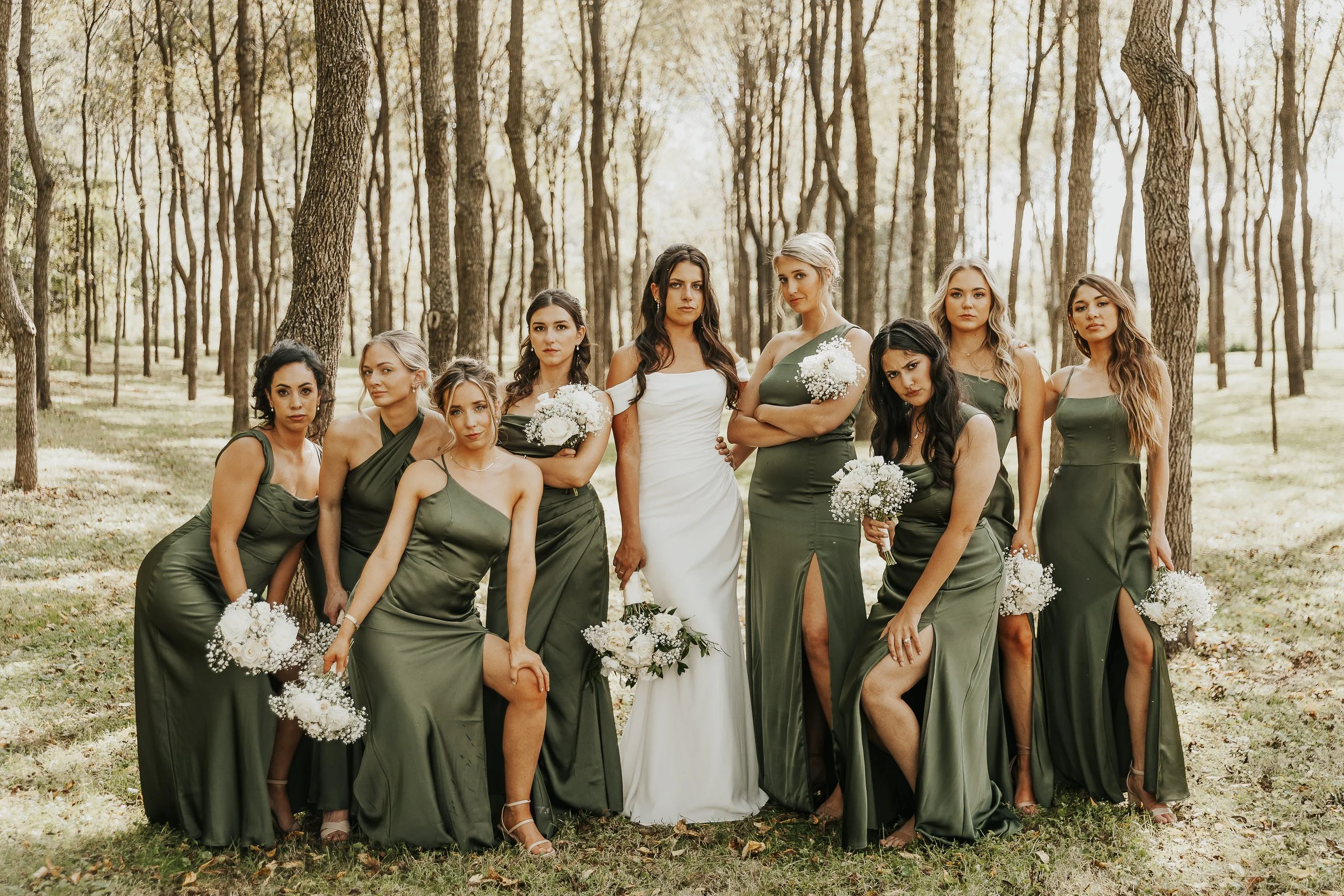 A bride and nine bridesmaids posing together outdoors in a wooded area during a wedding. The women are dressed in matching olive green dresses, and the bride is in a white gown holding a bouquet. The bridesmaids are also holding bouquets of white flo