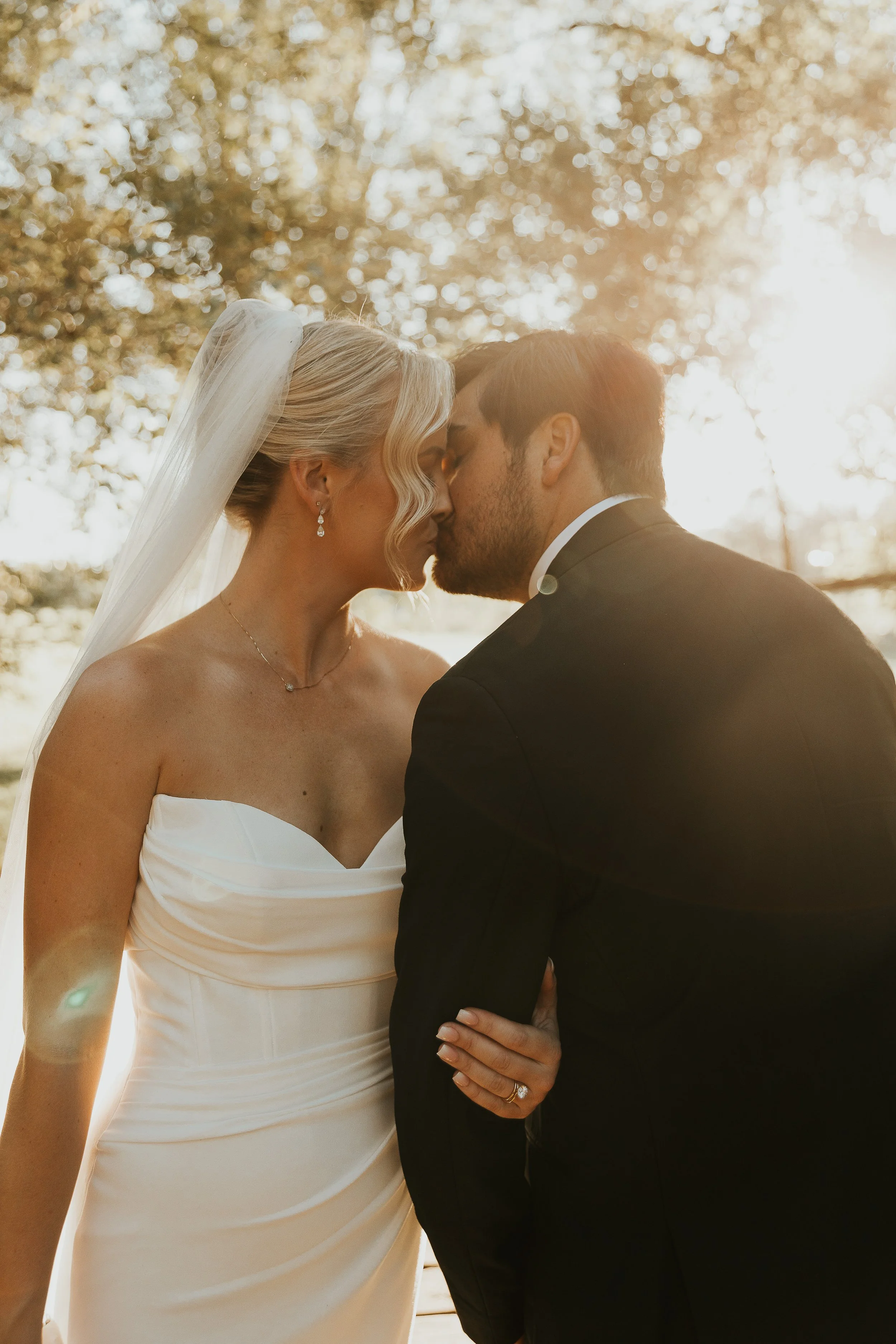 A bride and groom are kissing outdoors in sunlight, with trees in the background.