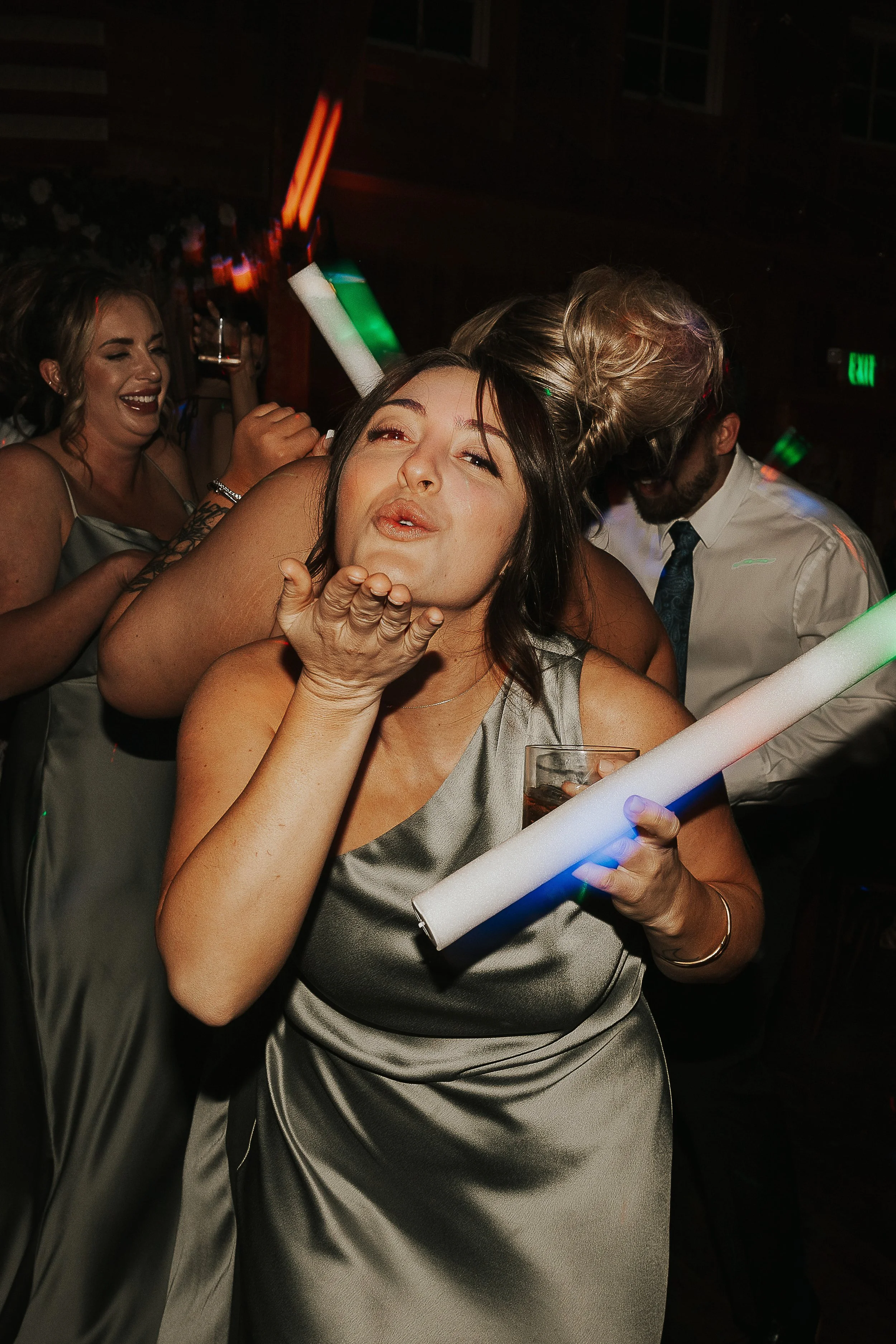 A woman blowing a kiss at a party or celebration, surrounded by friends, with party glow sticks and drinks, wearing a silver dress.
