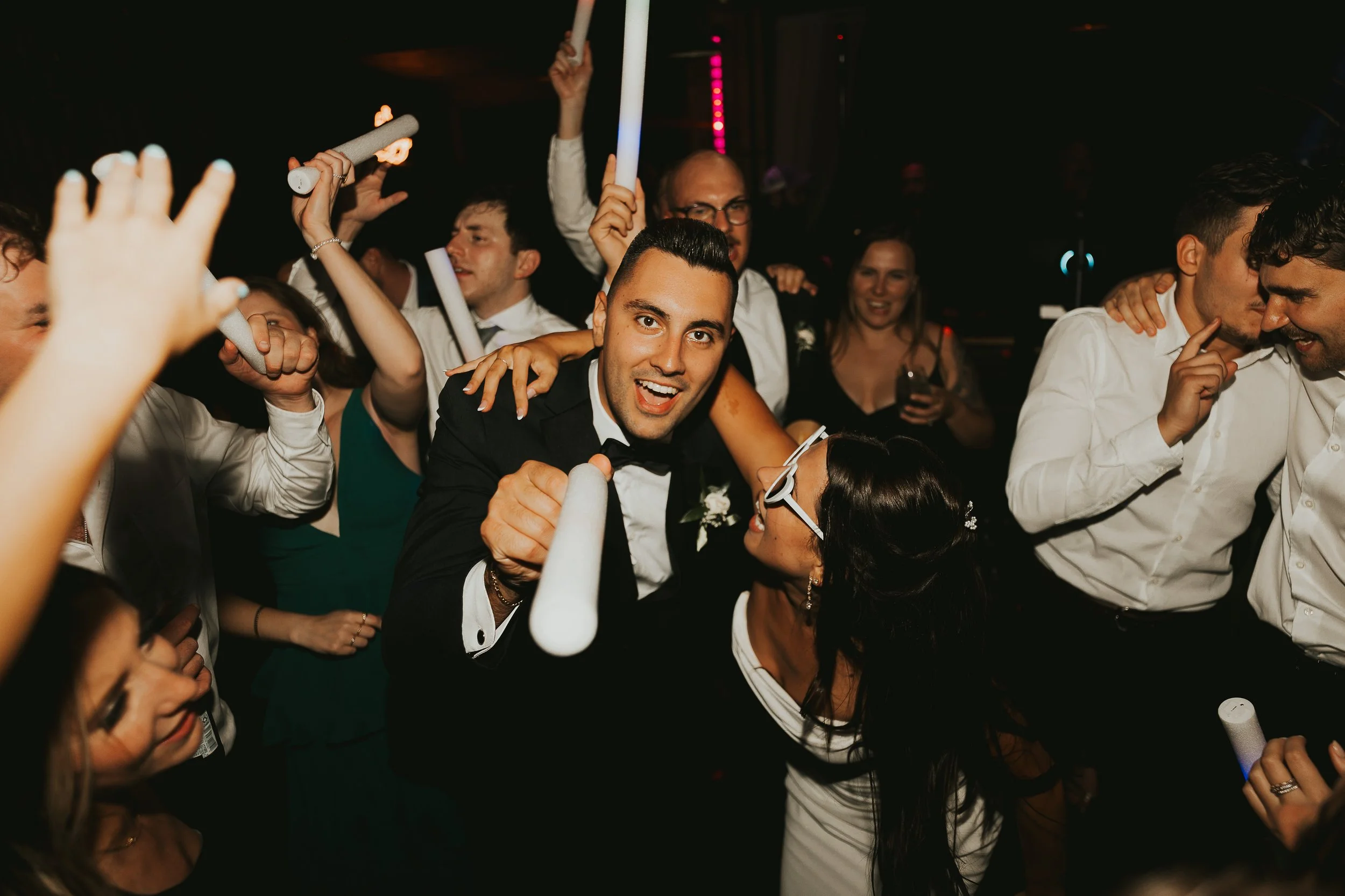 Group of people dancing and celebrating at a party with some holding rolled-up papers or glow sticks, smiling, and embracing each other in a dark setting with colorful lights.