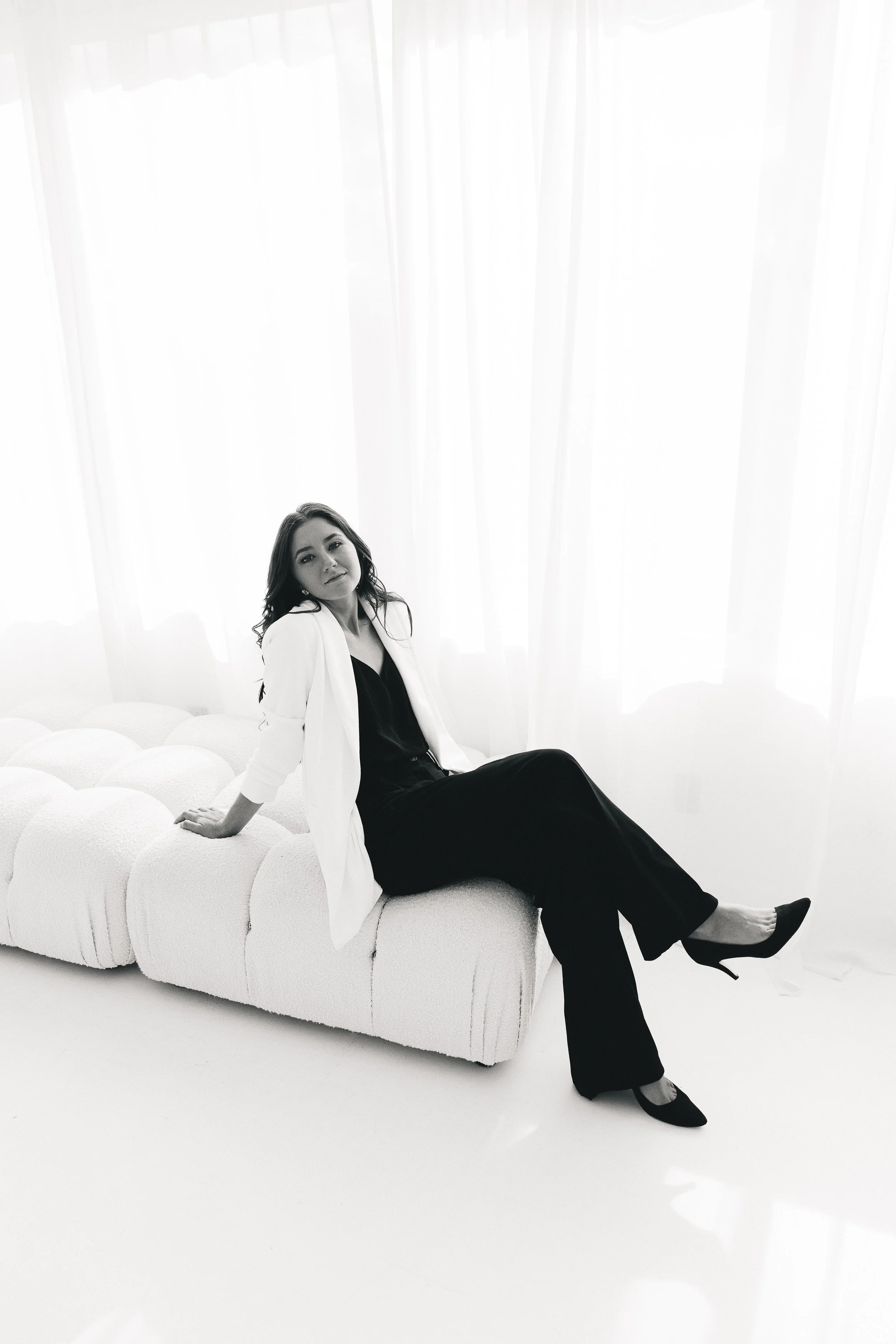 A photographer sitting on a modern white sofa in front of sheer curtains with soft natural light, wearing a white blazer, black top and black pants, with one leg crossed and wearing high heels.