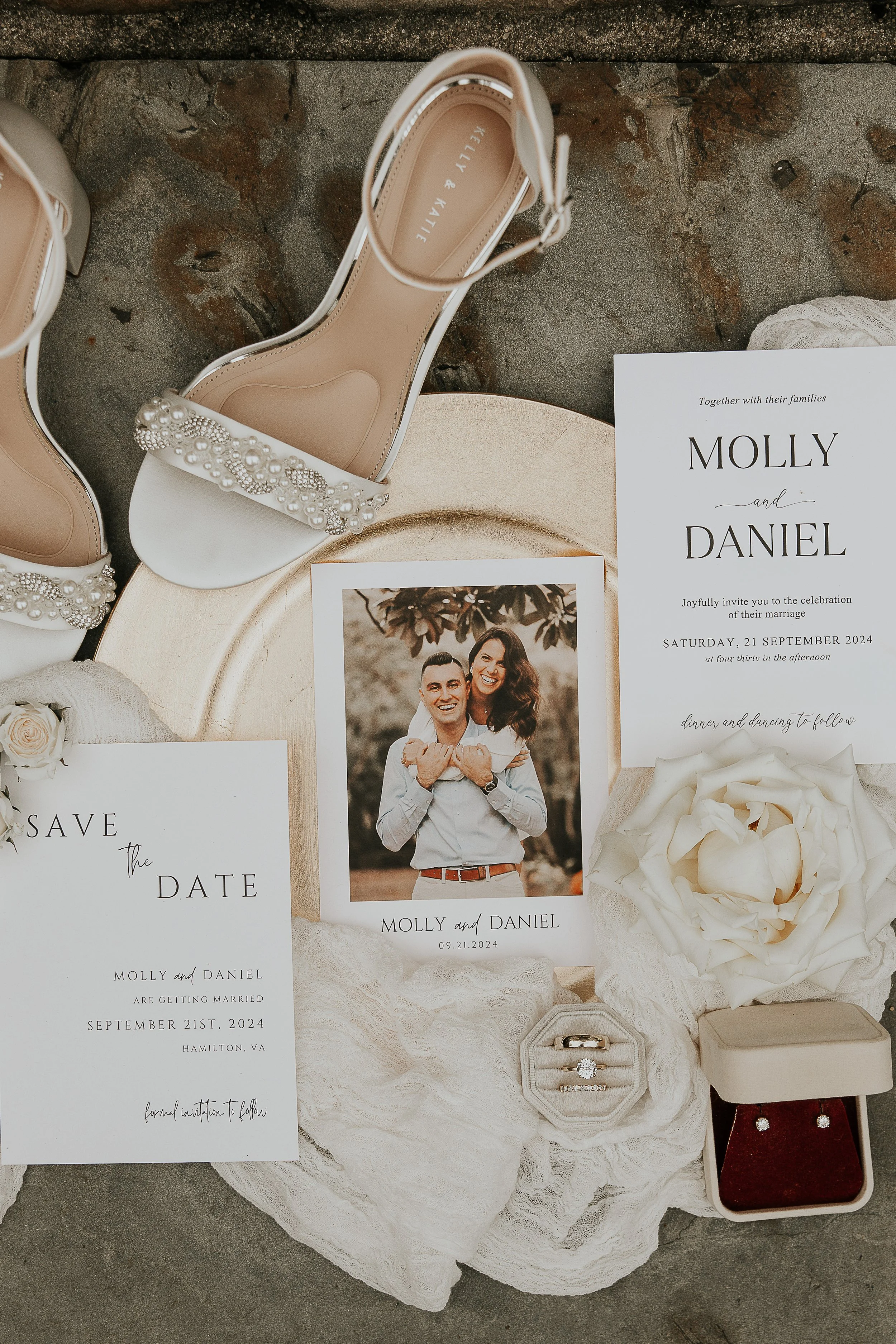 Wedding invitation cards, a photo of a smiling couple, pearl embellished white wedding shoes, a bouquet of white roses, a small jewelry box with rings, a red jewelry box, and other wedding accessories are displayed on a textured gray surface.
