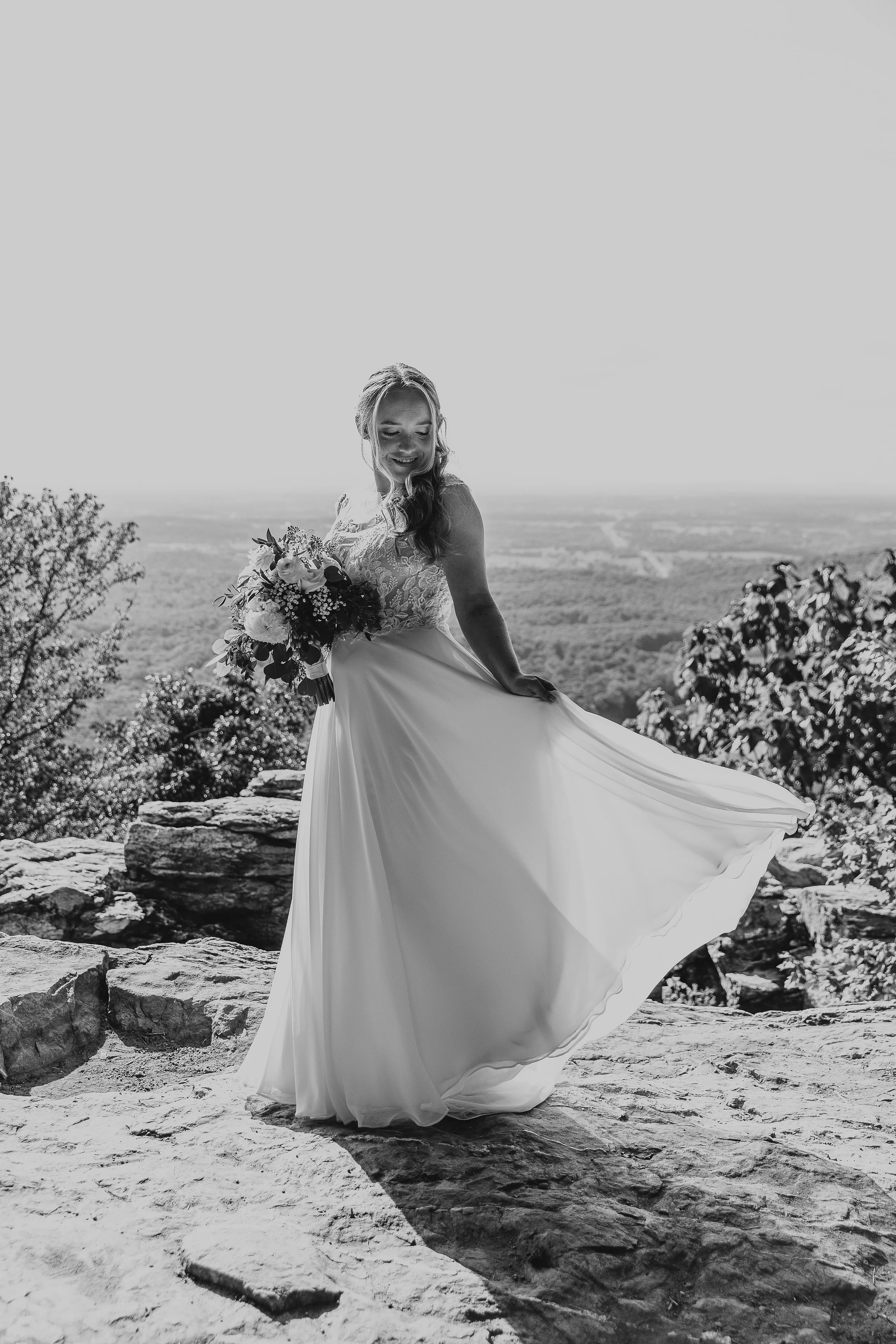 A black and white photo of a smiling elopement bride in a wedding dress holding a bouquet, standing outdoors with rocks and trees, overlooking a distant landscape.