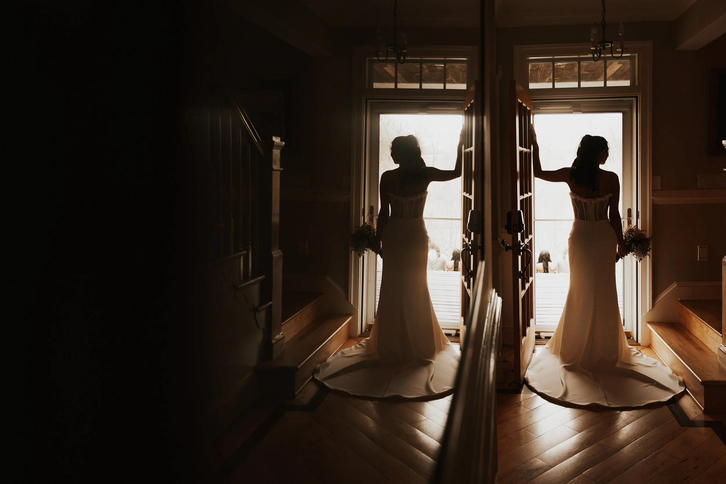 Two women in wedding dresses standing in front of open glass doors, holding bouquets, with light streaming in from outside.