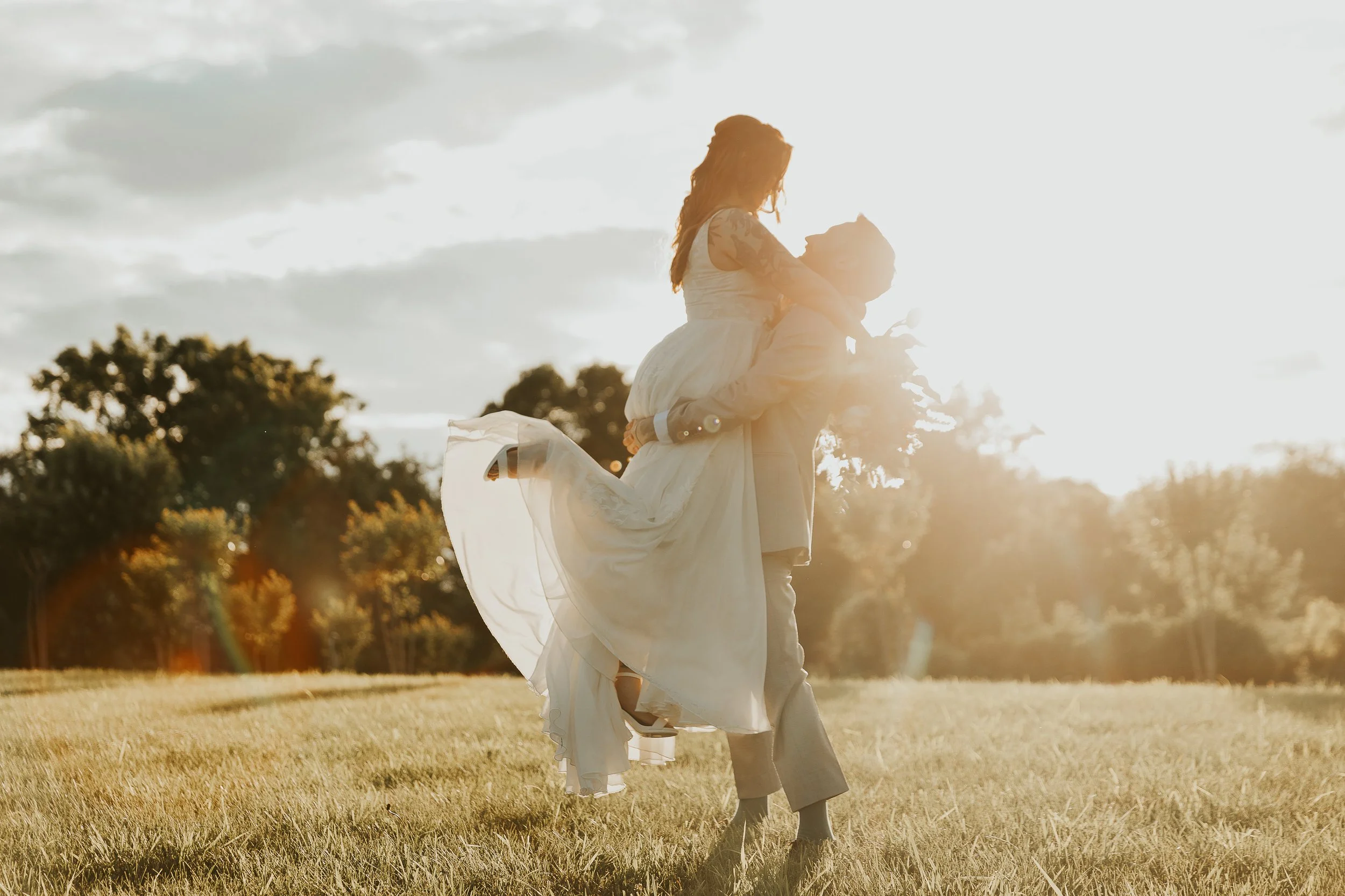 A couple dressed in wedding attire, with the woman sitting on the man's shoulders in a grassy field during sunset, smiling and enjoying the moment.