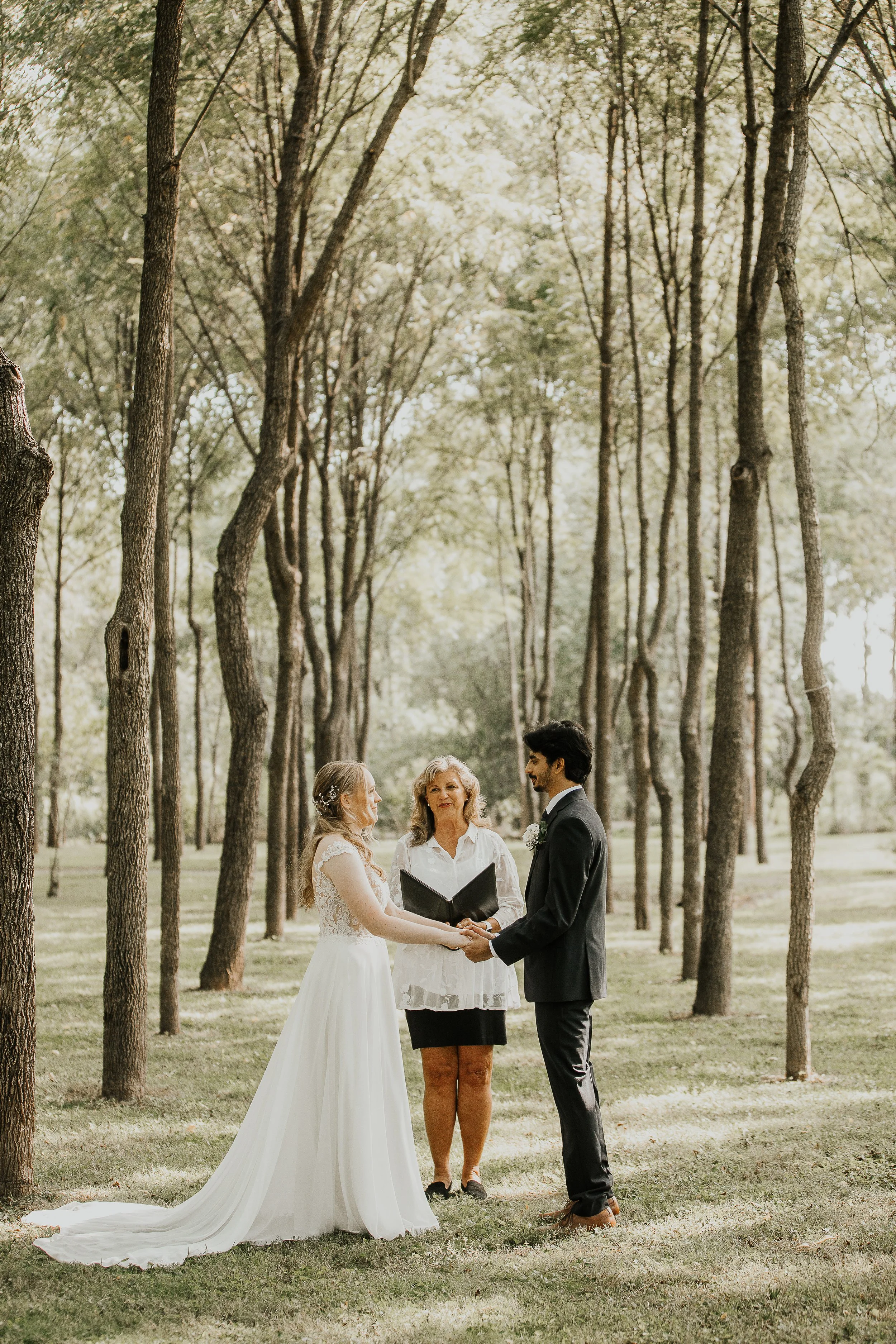 A couple getting married outdoors in a wooded area, holding hands and facing each other with an officiant standing in the background, all surrounded by tall trees and sunlight filtering through the leaves.