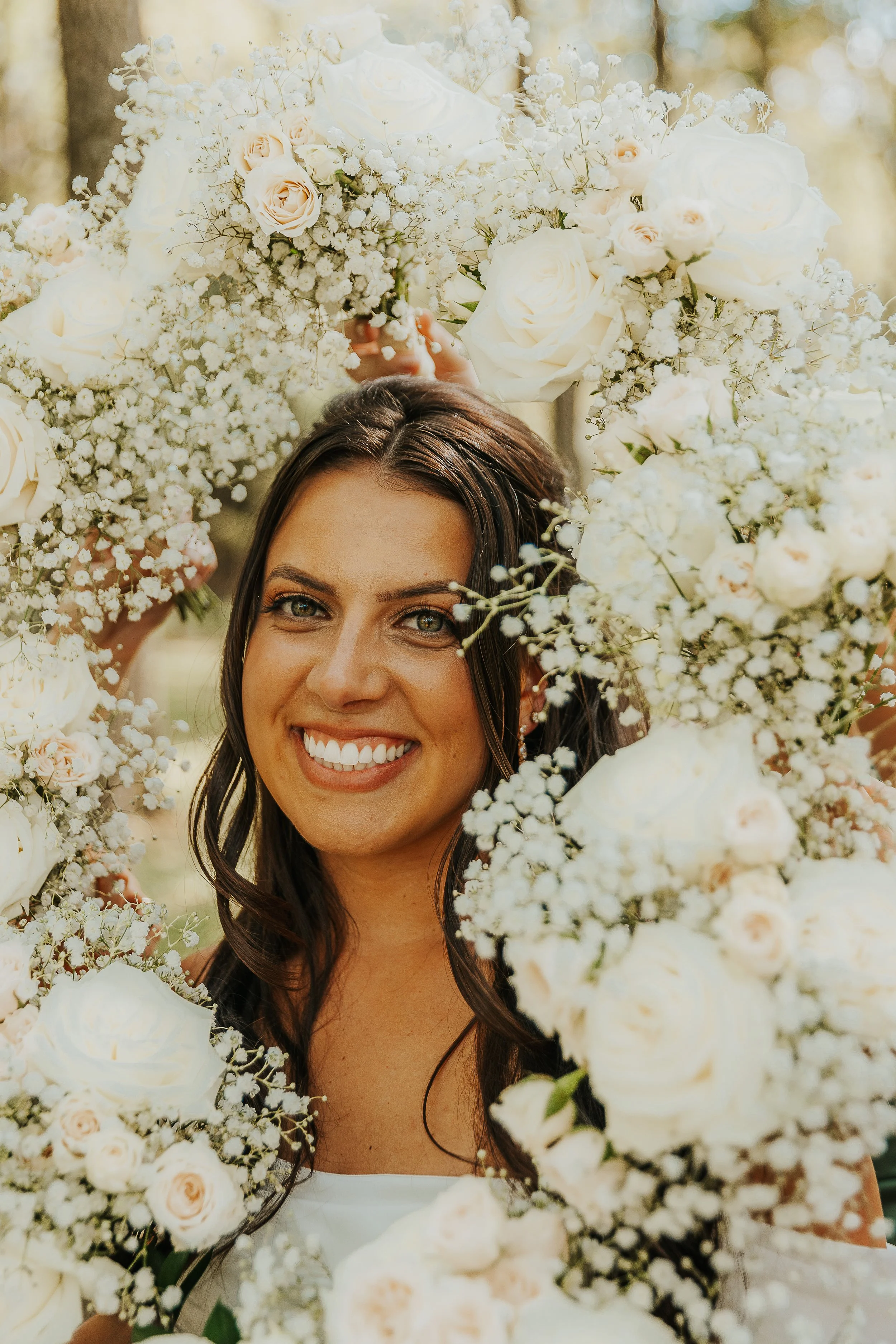 A woman with long dark hair and a big smile surrounded by a large arrangement of white and pale pink roses, baby's breath, and greenery, outdoors in natural light.