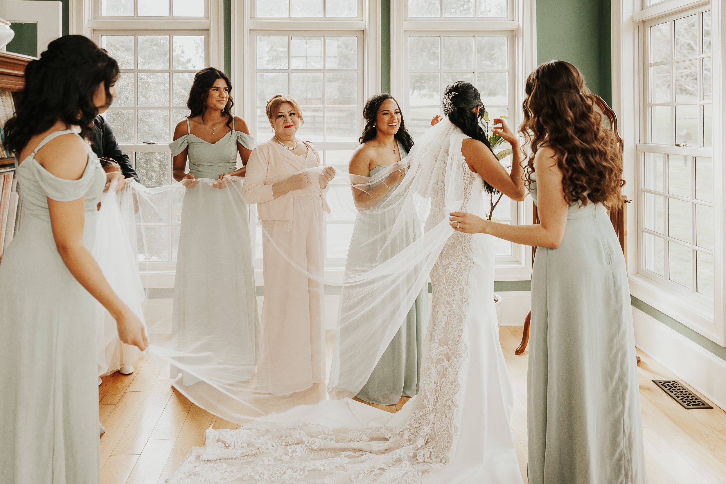 A bride wearing a lace wedding dress holding a flower, standing face-to-face with another woman in a light blue dress while friends and family gather around. The scene takes place in a bright room with large windows.