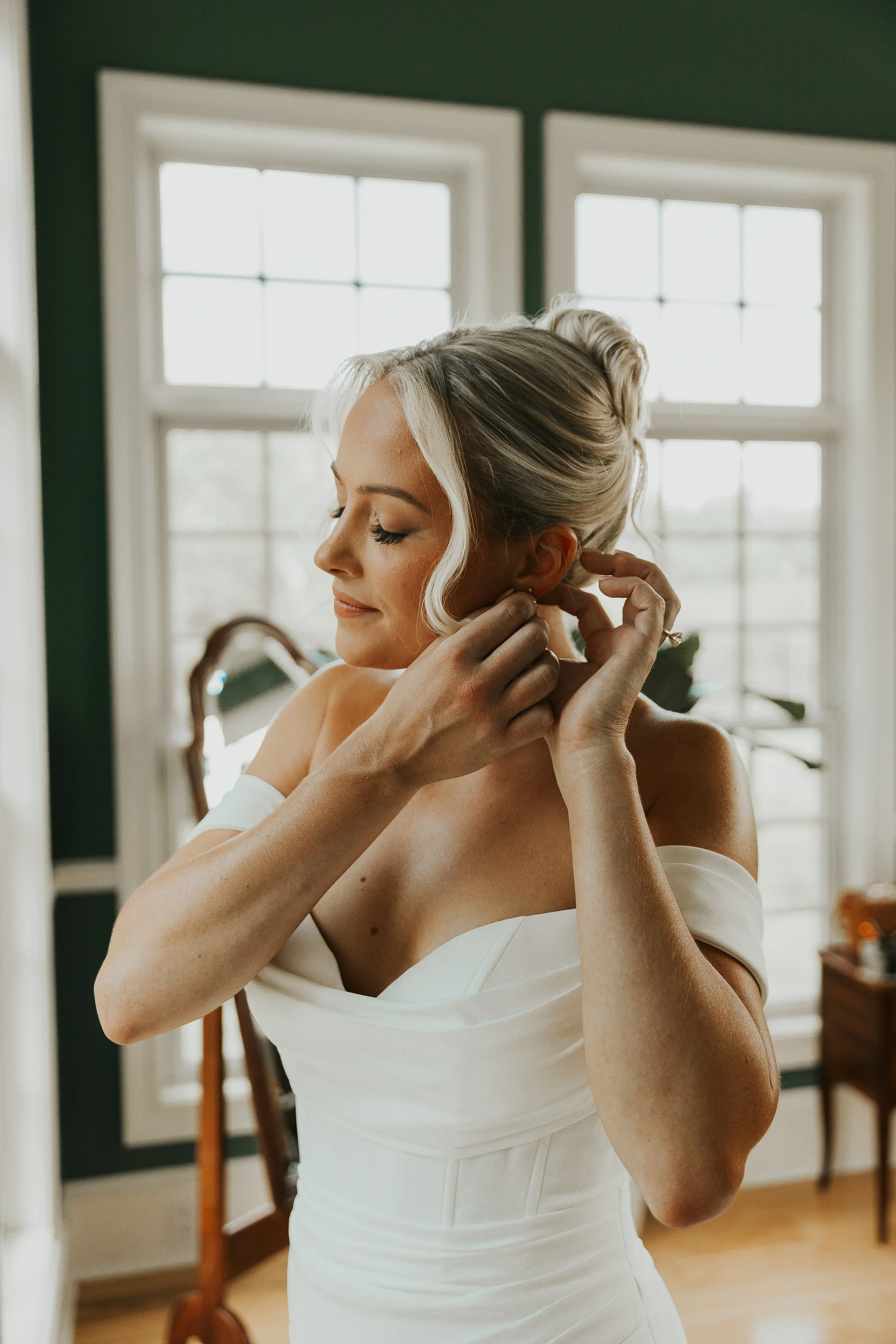 A woman with blonde hair styled in an updo, wearing a white off-the-shoulder dress, adjusts her earring in a room with a large window and green walls.