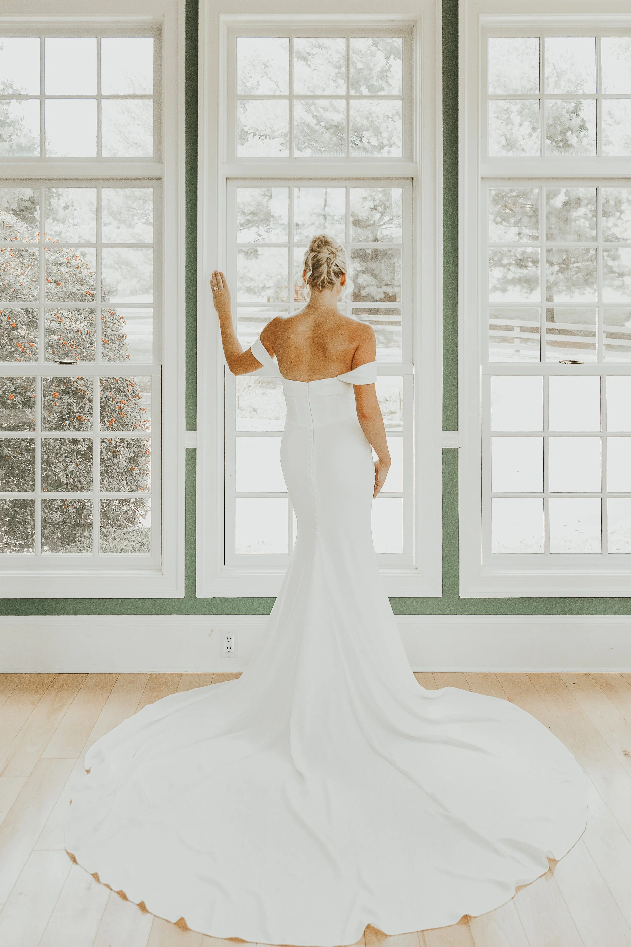 A woman in a white strapless wedding dress standing by large windows, looking outside, with her back to the camera.