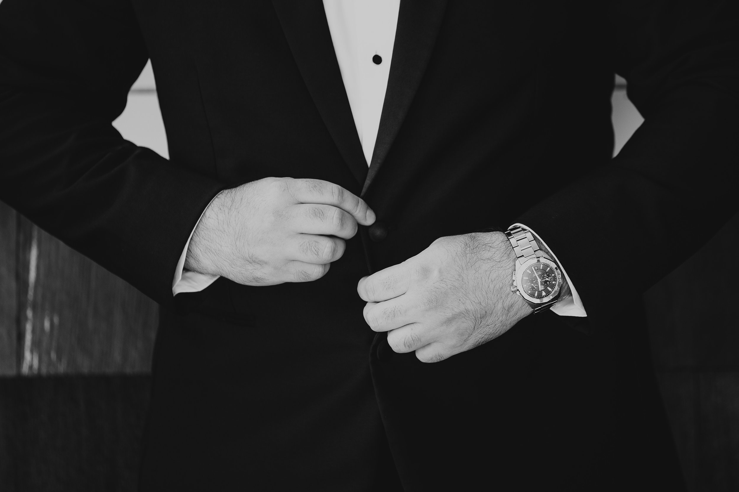Close-up of a man in a tuxedo buttoning his jacket, wearing a wristwatch.
