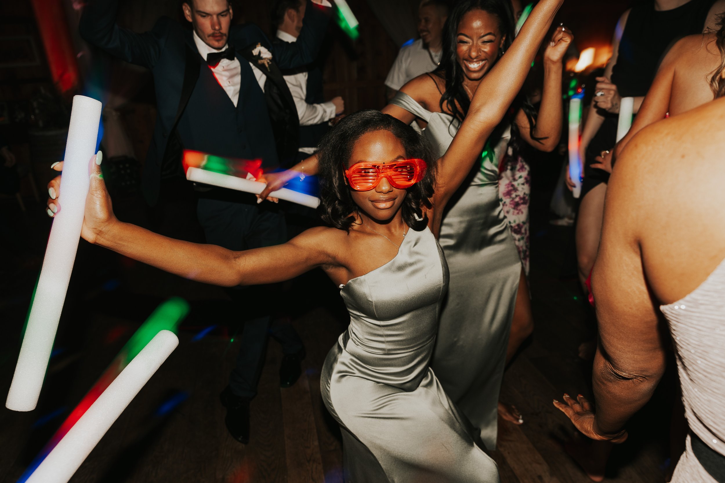 Group of people dancing at a party, with a woman in a silver dress and red glasses in the foreground, and a man in a tuxedo in the background.