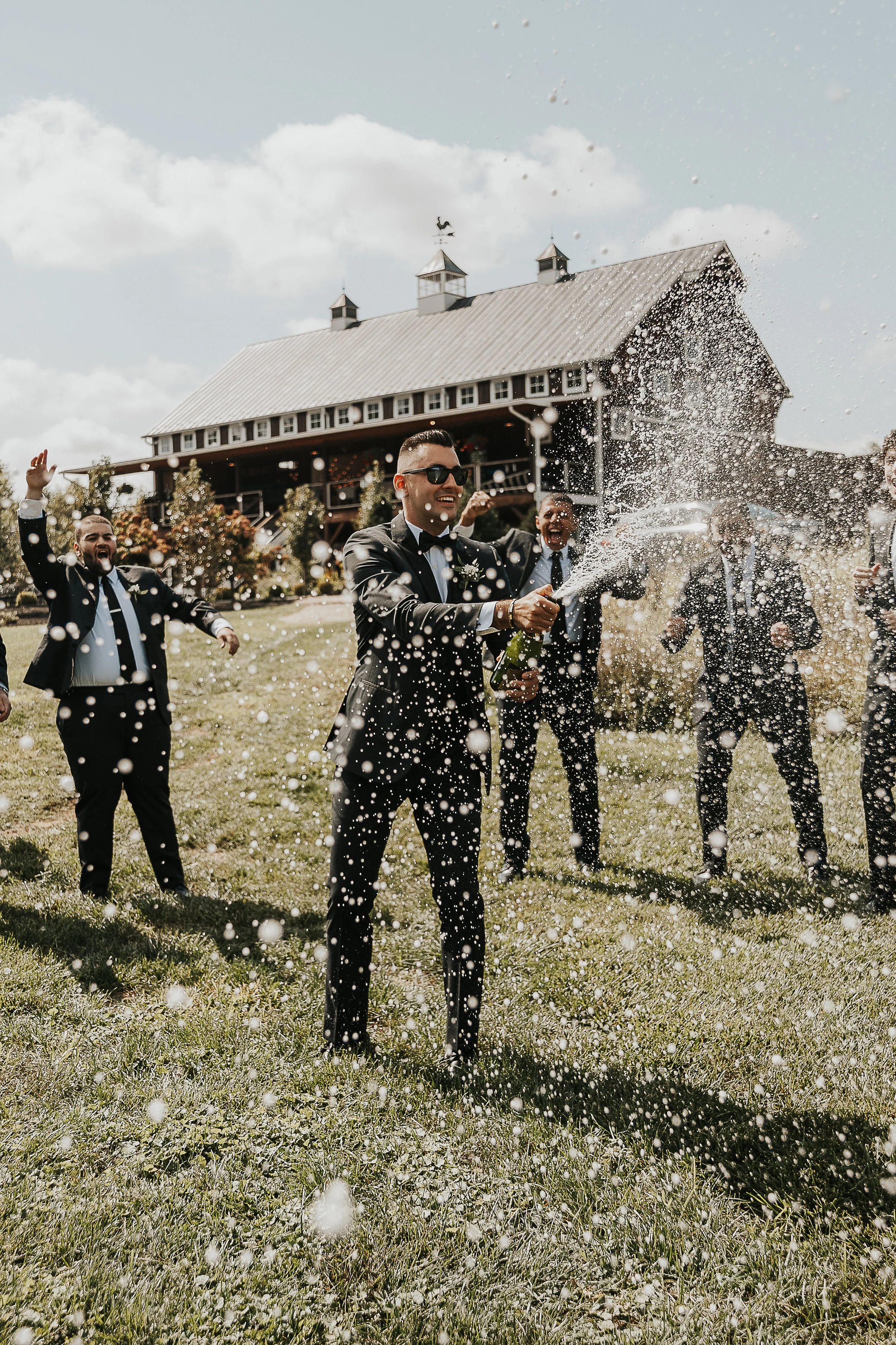 Group of men dressed in tuxedos celebrating outdoors, with a man spraying champagne, in front of a large barn-style building on a sunny day.