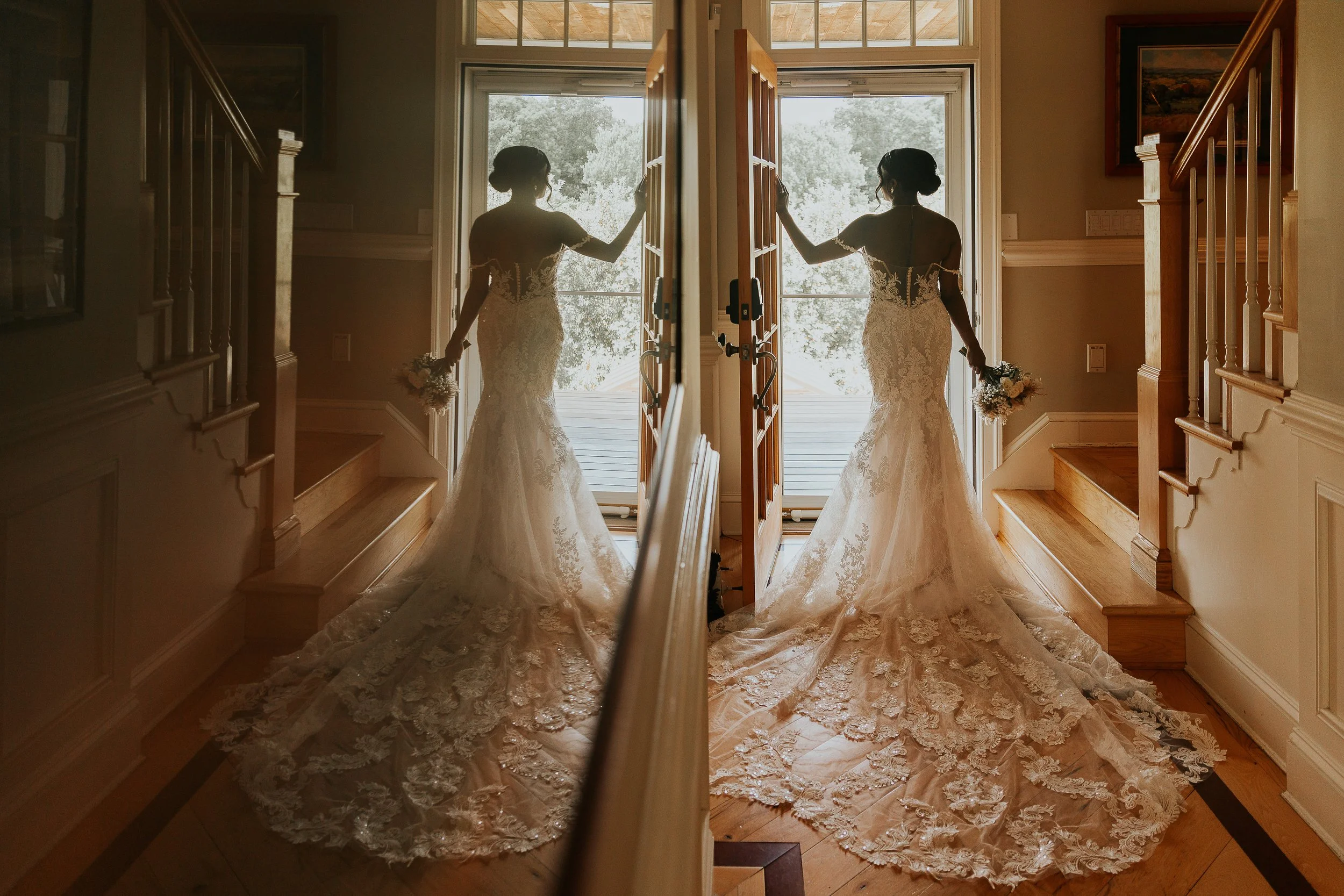 A bride in a lace wedding gown is standing in front of a large window, holding a bouquet, with her reflection visible in a mirror that also reflects the window and trees outside, inside a well-lit room with wooden stairs and framed artwork.