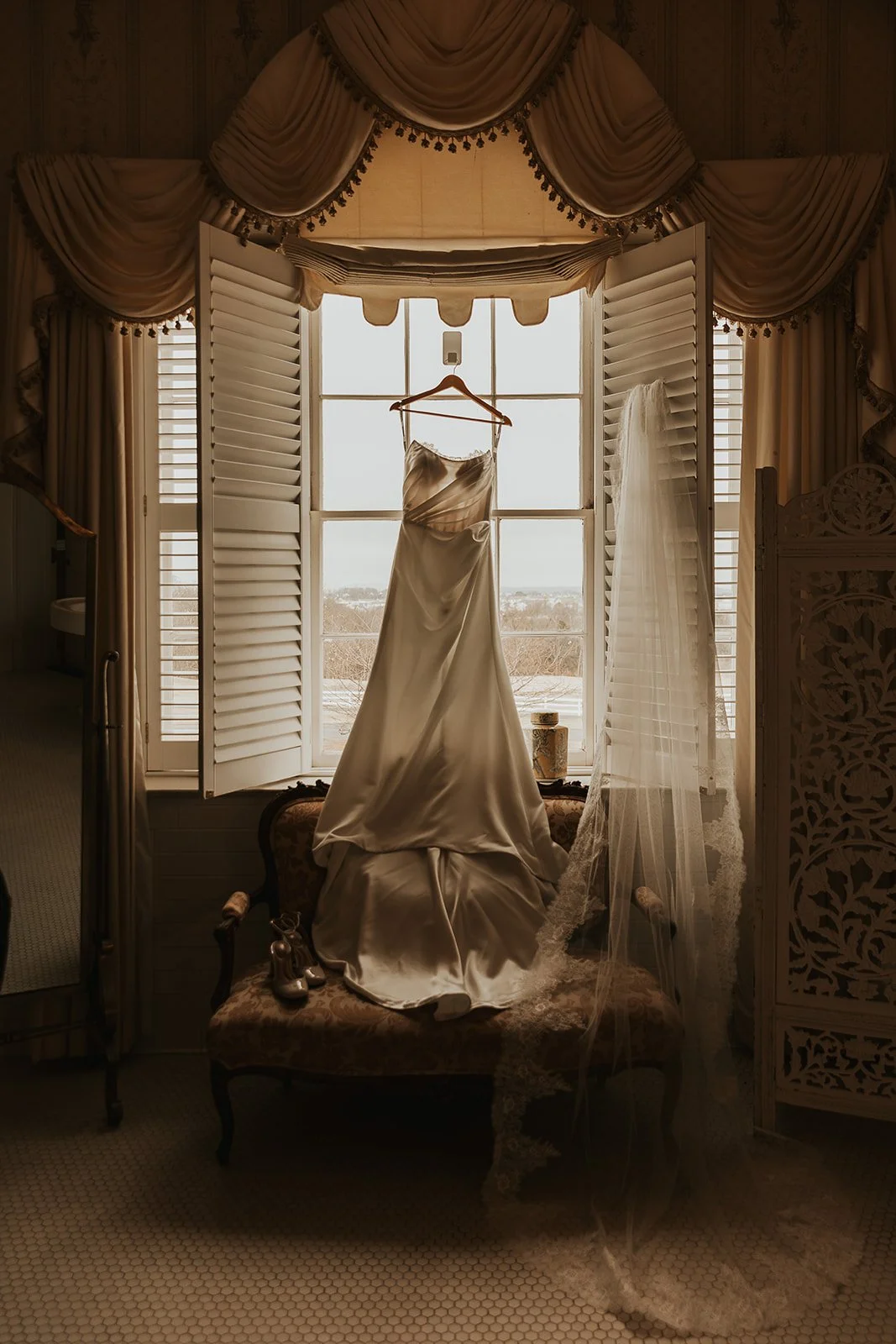 A satin wedding dress hanging on a hanger in front of a window with open shutters, a veil draped over a chair, and bridal shoes on the chair, with natural light illuminating the scene.
