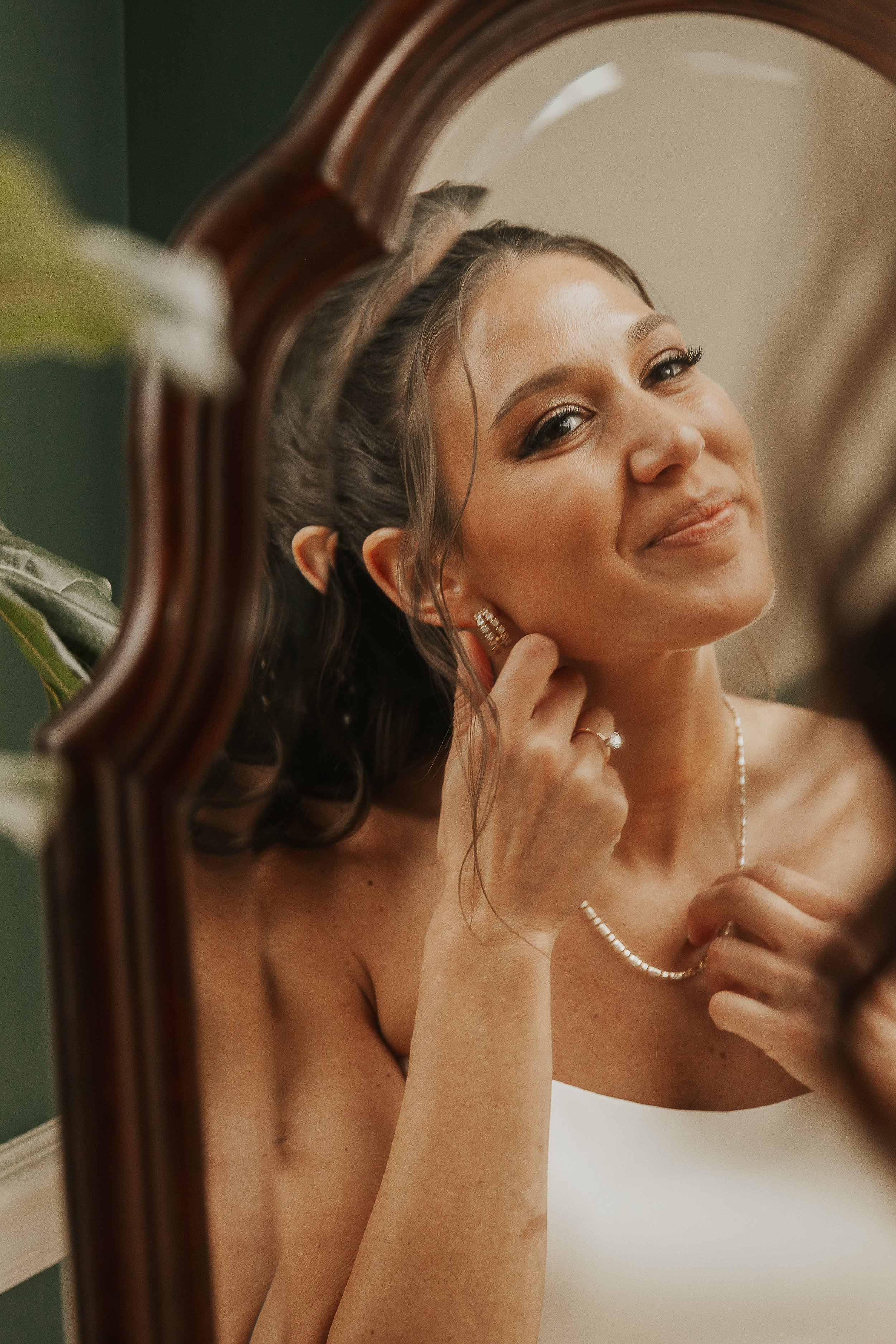 A woman looking in a mirror, adjusting her earring, with jewelry including a necklace and rings, in a room with a green wall.