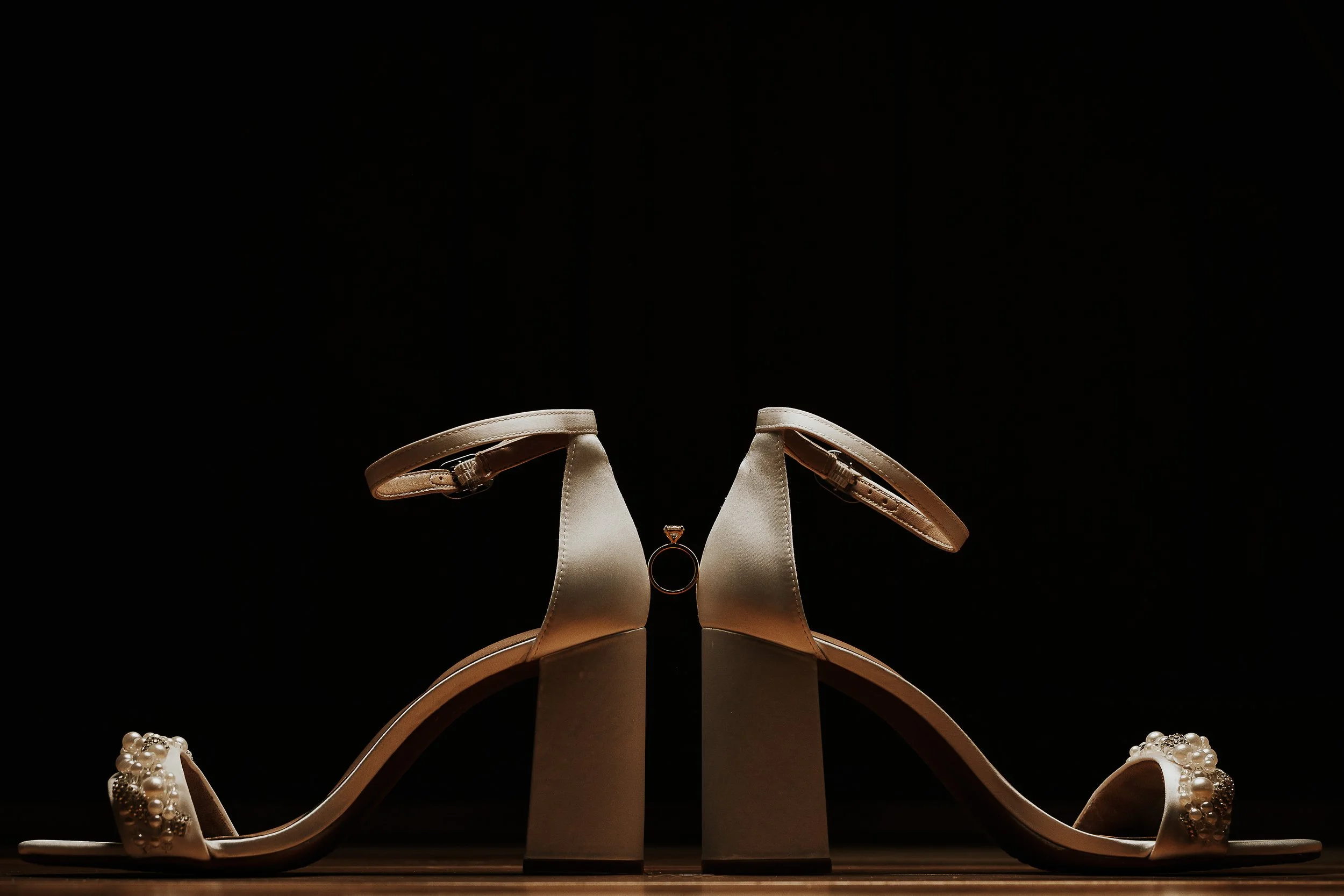 A pair of elegant high-heeled shoes with ankle straps, displayed on a stand against a black background.