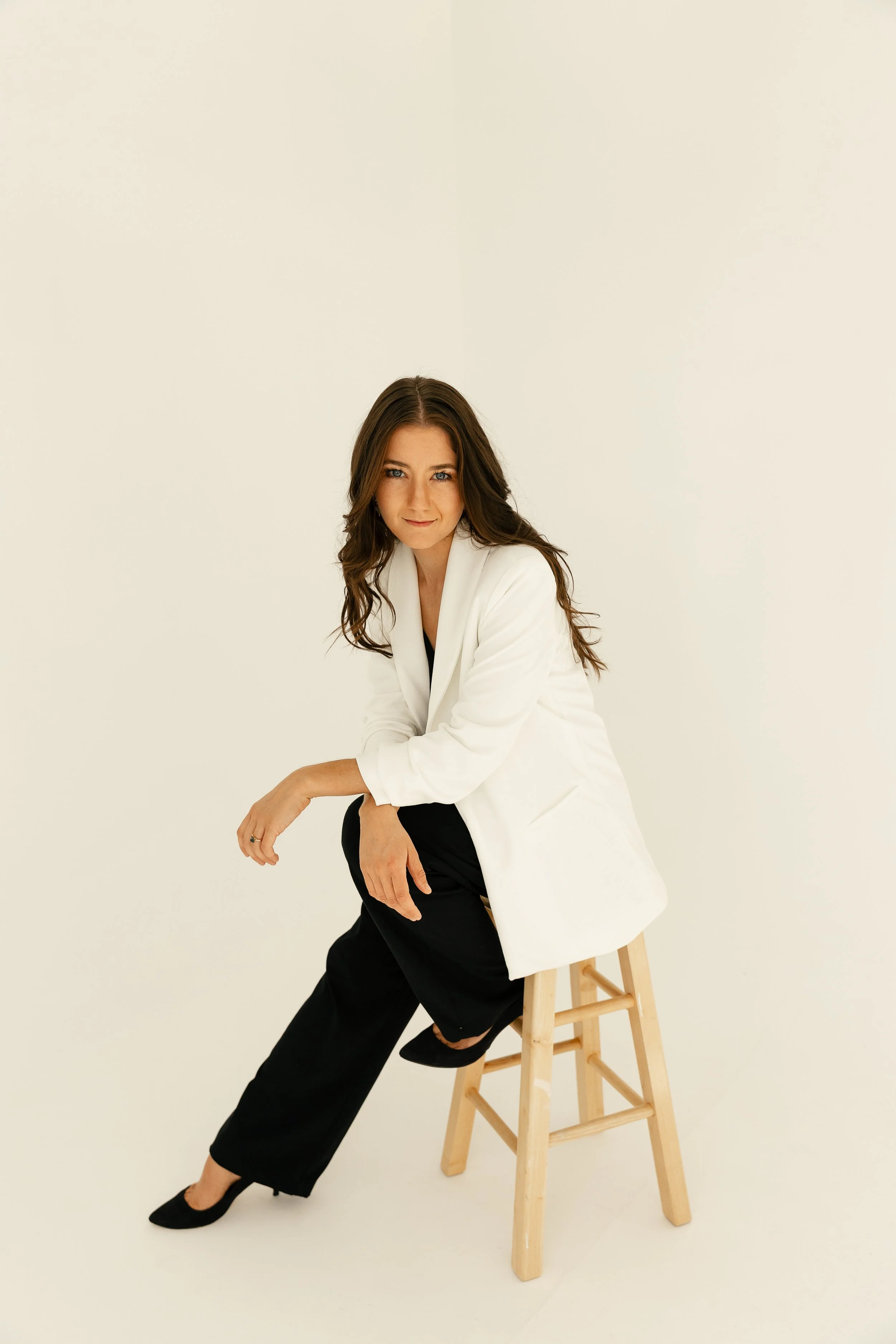 A photographer with long brown hair and blue eyes sitting on a wooden stool, wearing a white blazer, black ripped pants, and black heels, against a plain off-white background.