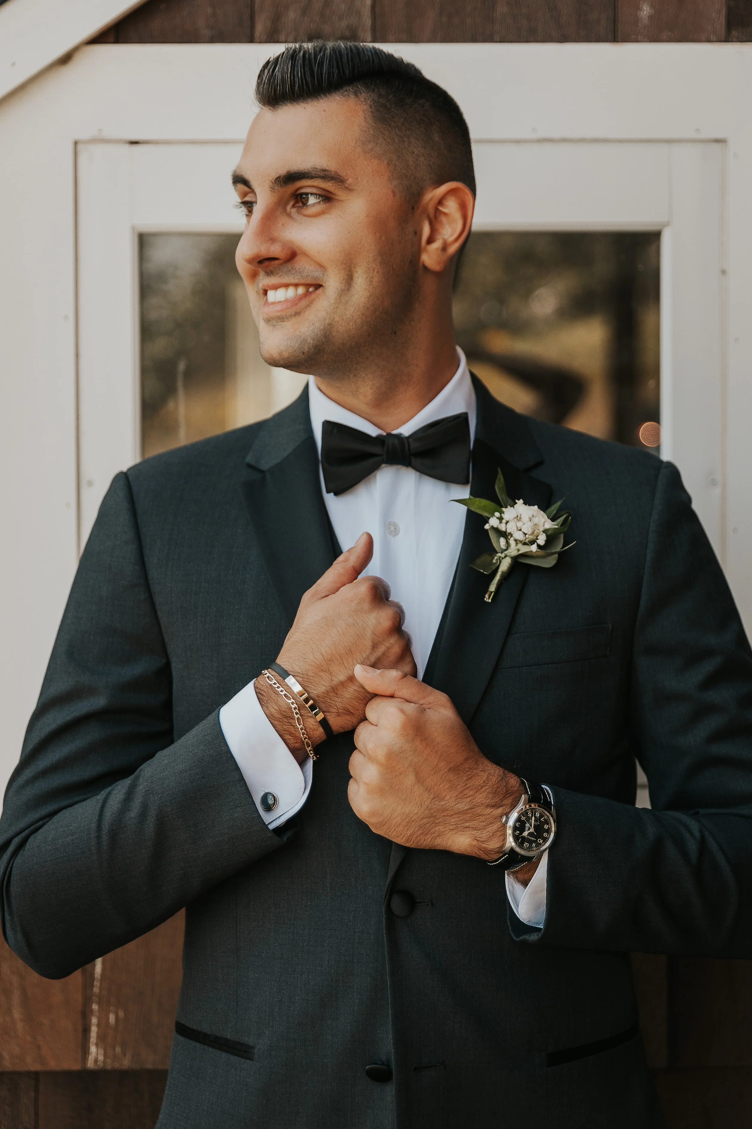 A man dressed in a black tuxedo with a white shirt and black bow tie, standing in front of a wooden background. He is smiling and looking to his left, adjusting his jacket with his right hand. He is wearing a wristwatch on his left wrist and a bracel