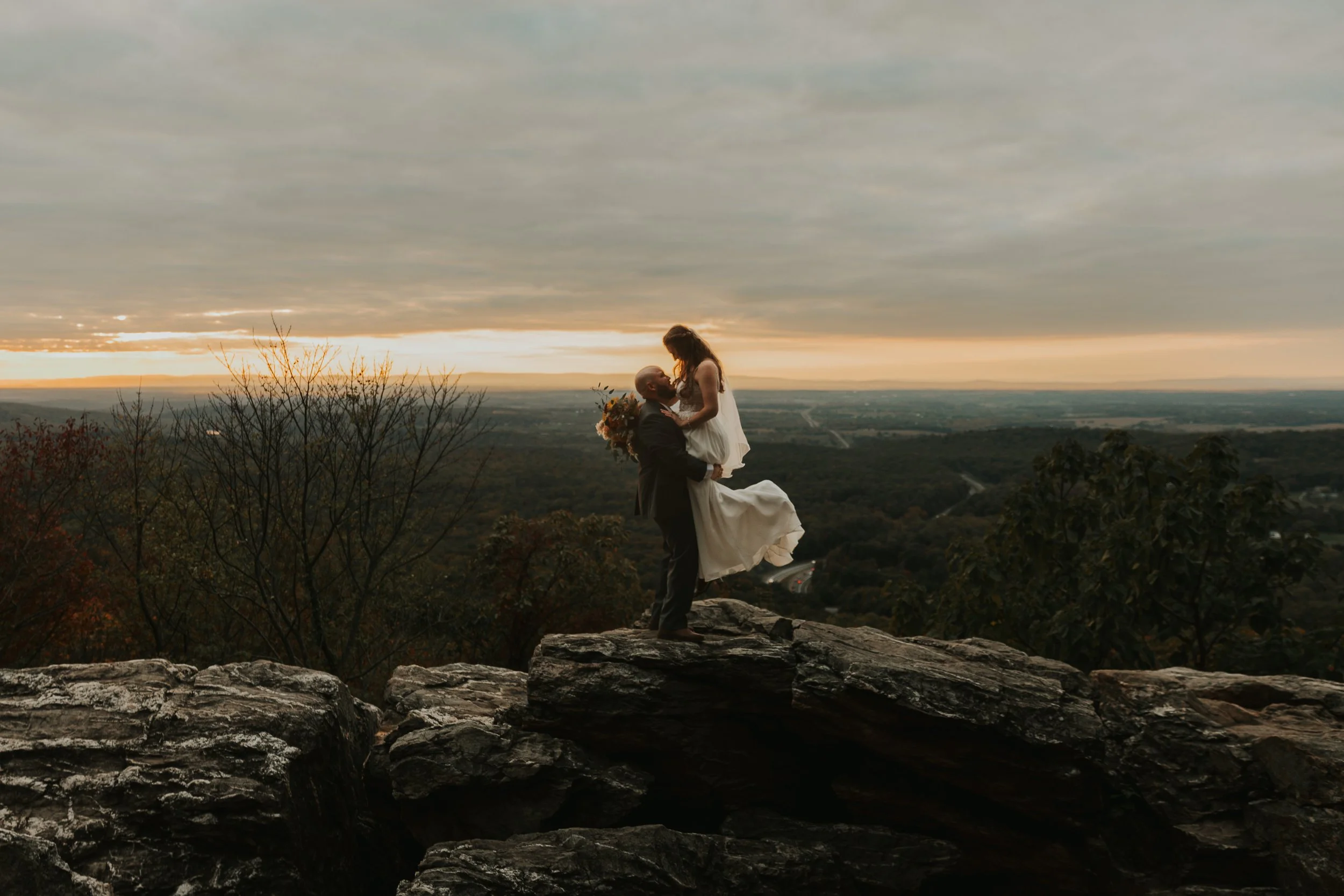 A man lifts a woman wearing a white dress in a scenic elopement outdoor location during sunset, with a panoramic view of the landscape and cloudy sky in the background.