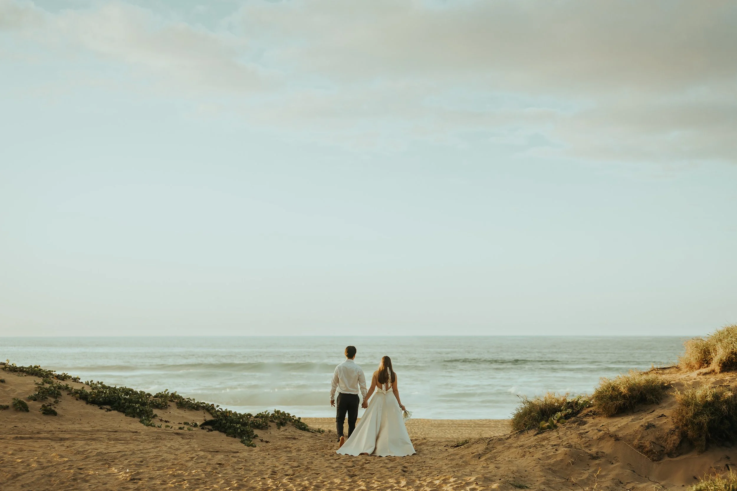 A destination wedding bride and groom walking hand in hand toward the ocean on a sandy beach, with sandy dunes and sparse vegetation on either side under a partly cloudy sky.
