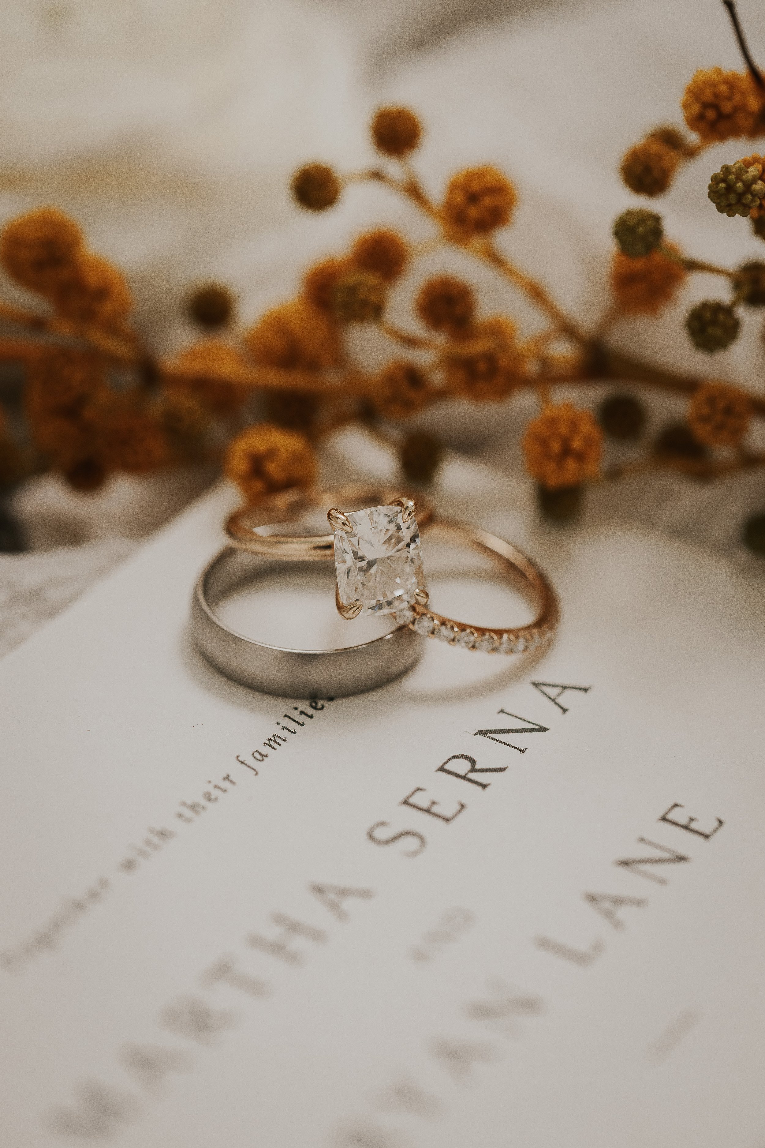 A close-up of two rings, one with a large square-cut gemstone and the other a band with small diamonds, placed on an elegant invitation with cursive writing. In the background, yellow and orange floral accents are blurred.