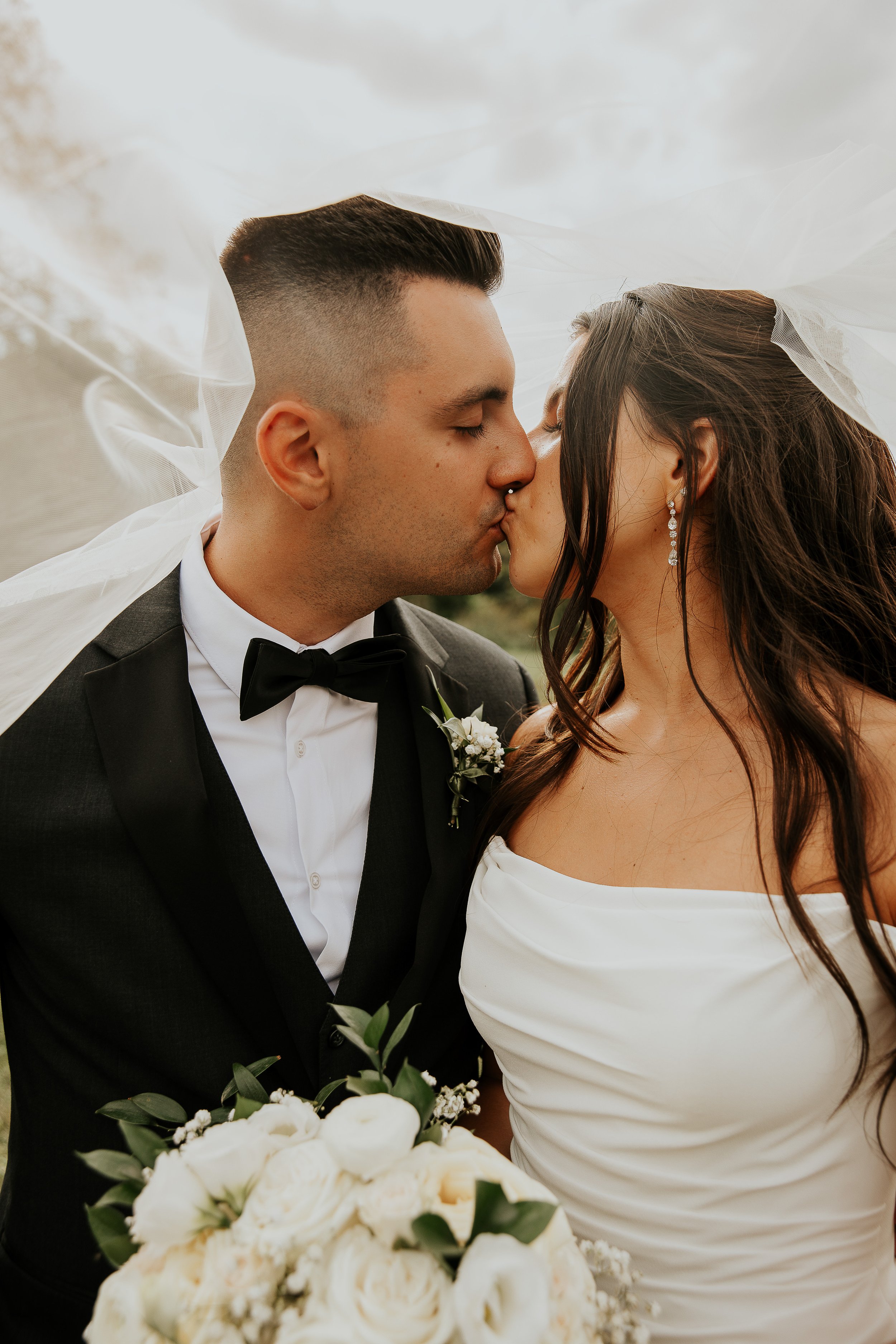 A bride and groom kissing on their wedding day, with the bride wearing a white dress and a veil, and the groom dressed in a tuxedo with a bowtie, holding a bouquet of white flowers.