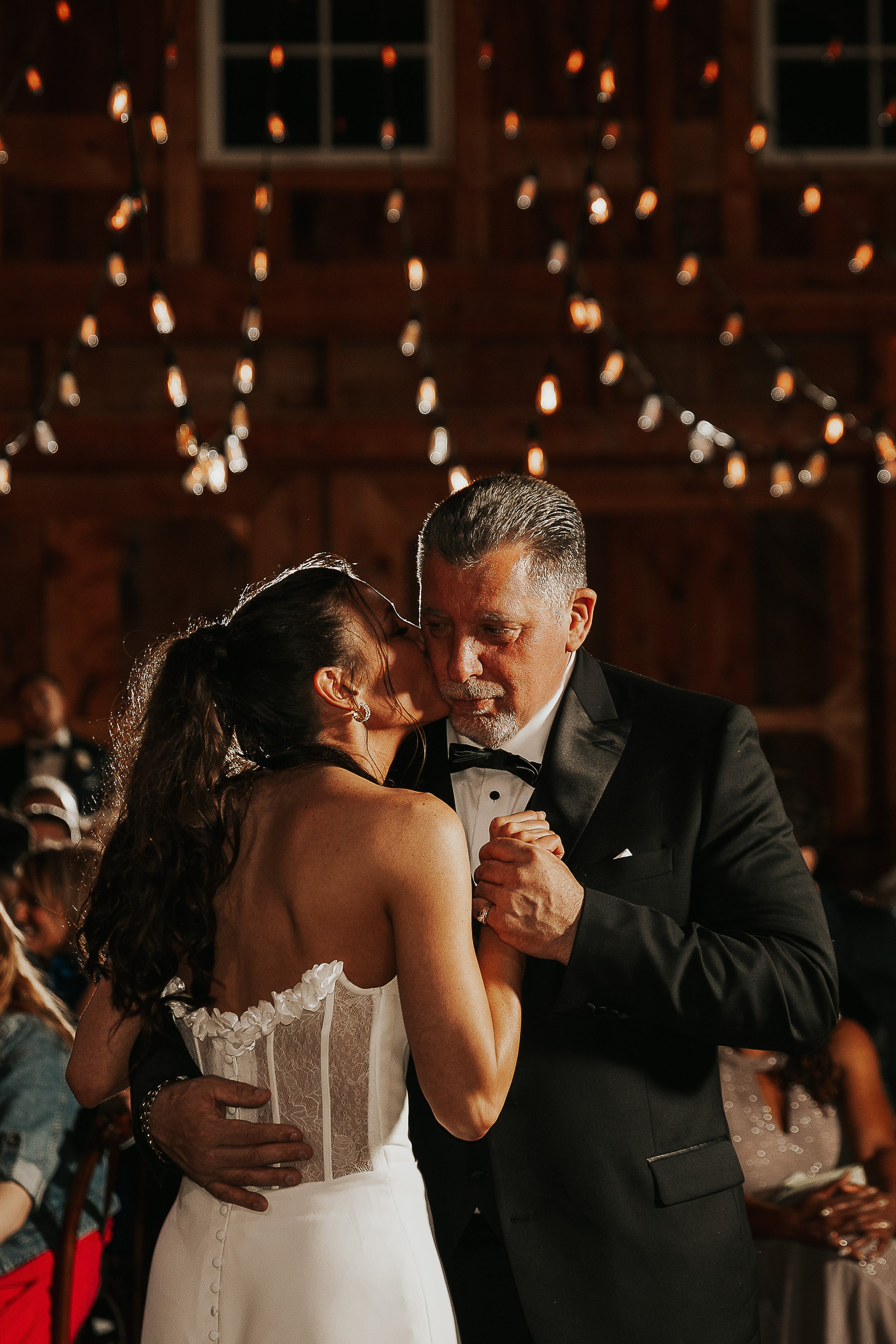 A bride and groom sharing a dance at their wedding reception. The bride is wearing a white dress with lace details, and the groom is dressed in a black tuxedo. They are holding hands and looking into each other's eyes. The background features warm li