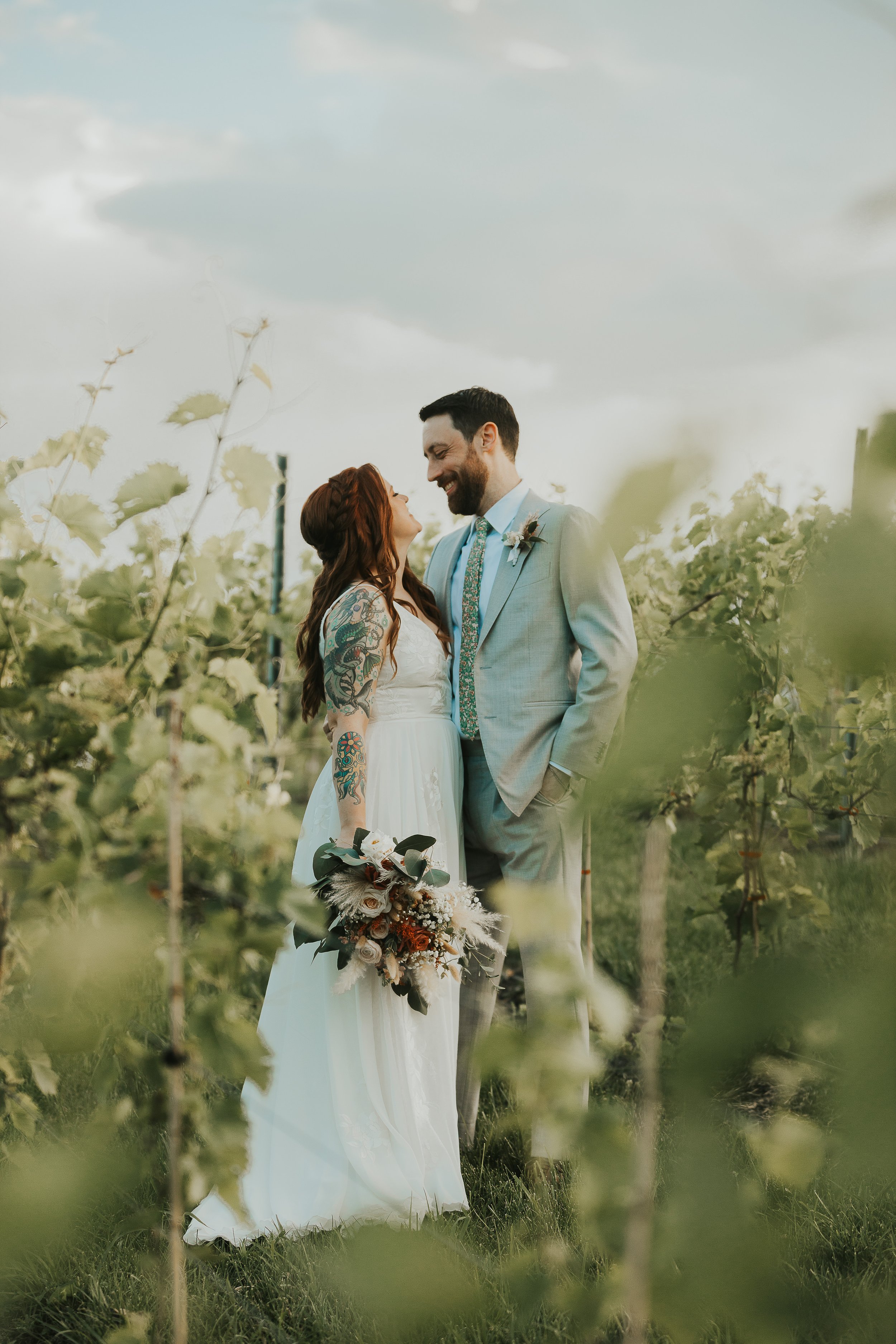 A bride and groom in a vineyard, smiling and looking at each other, surrounded by green grapevines and cloudy sky.