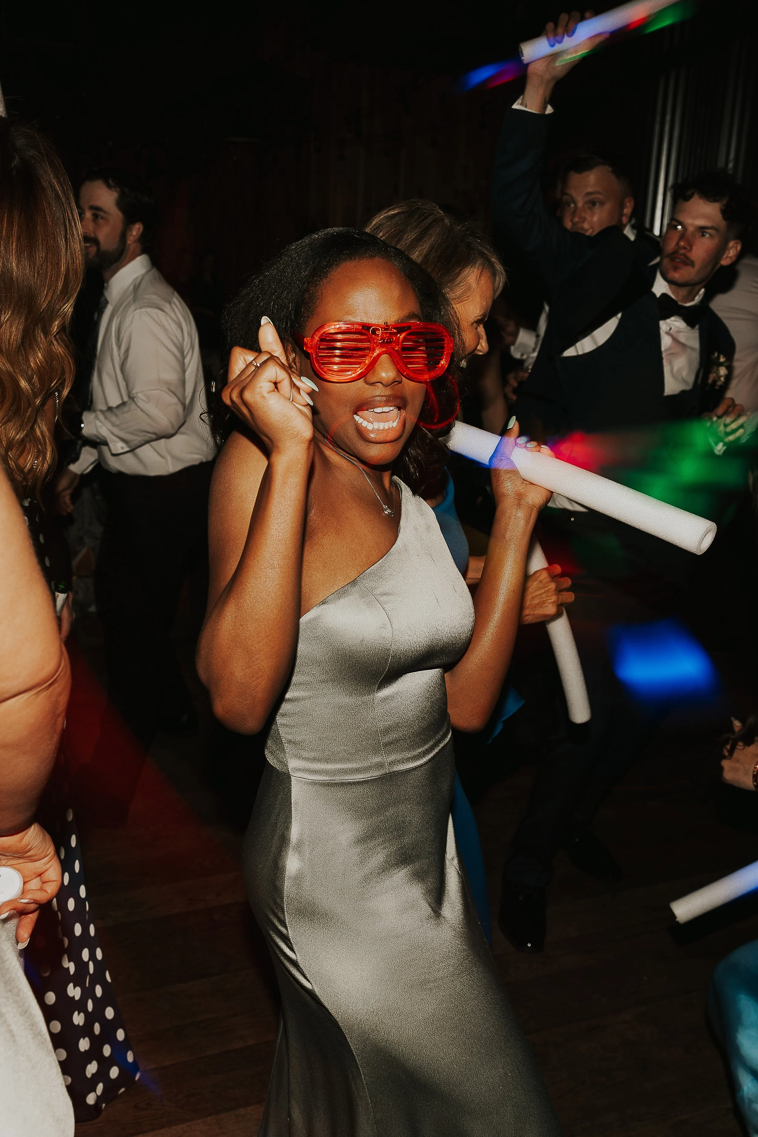 A woman wearing red shutter shades and a silver dress at a party, holding a glow stick, with people dancing in the background.