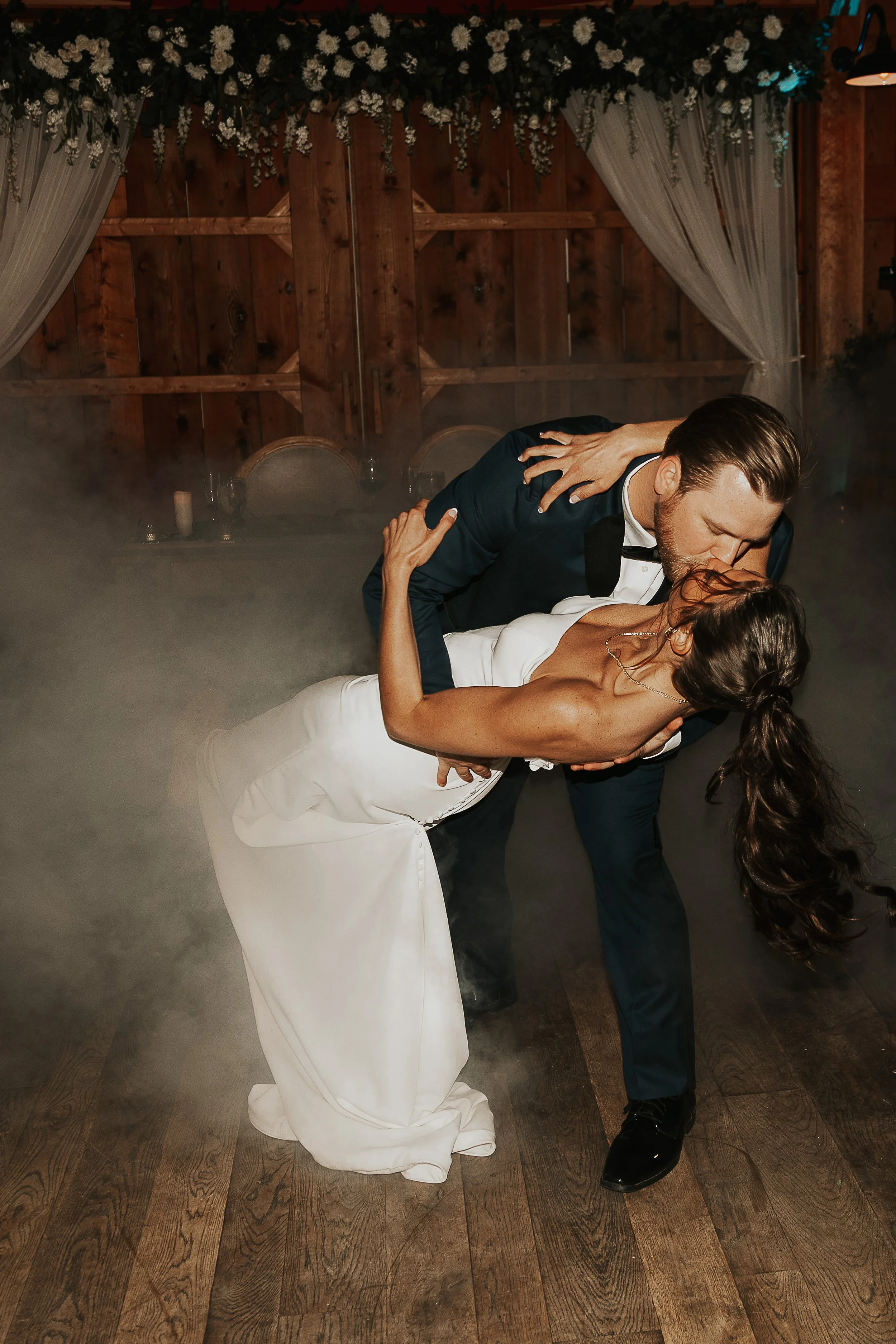 A man and woman dancing closely at a wedding reception, with the man dipping the woman backward. The woman wears a white wedding gown and the man a dark suit with a bow tie. The background features wood-paneled walls and floral decorations.