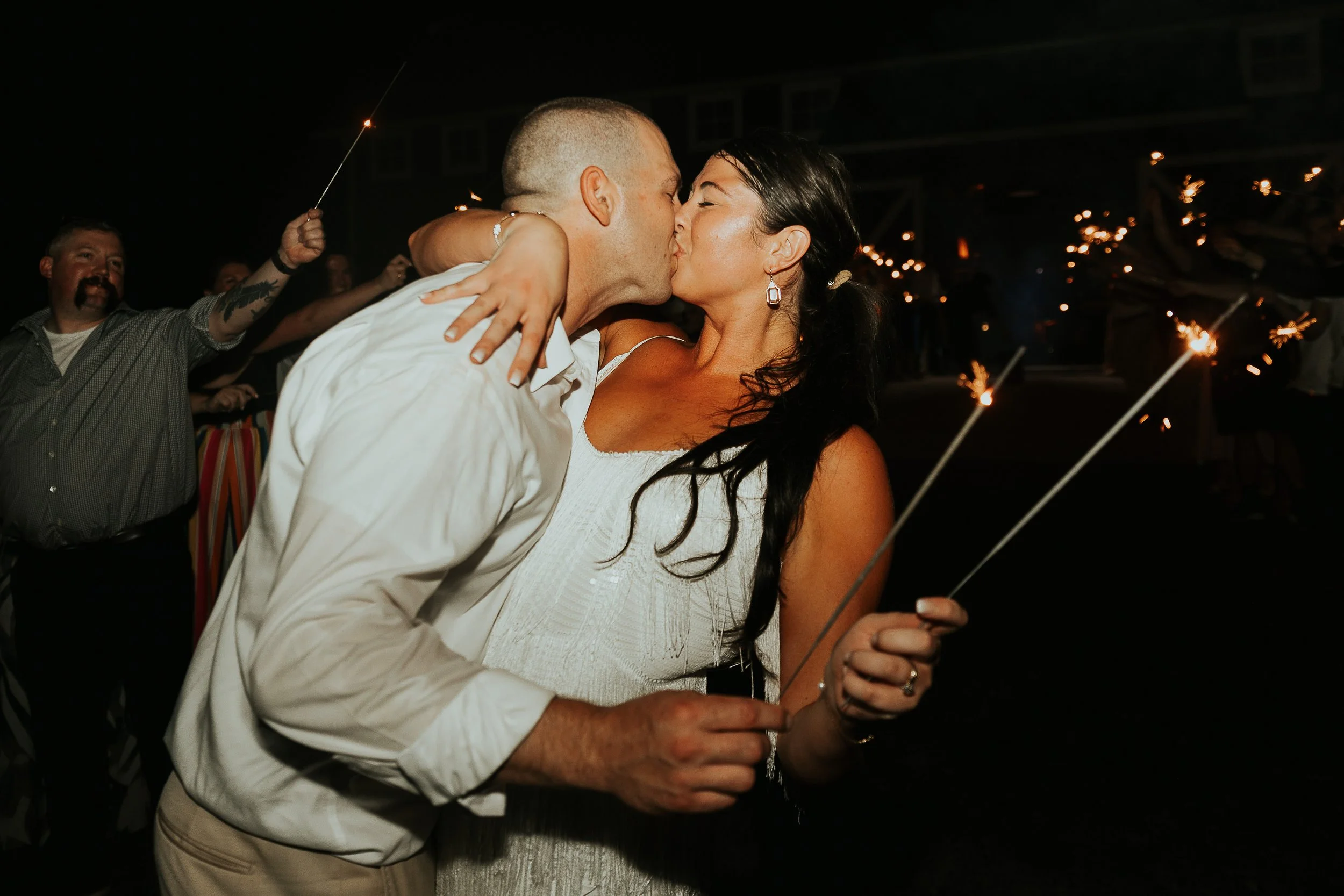 A couple is kissing at night, holding sparklers, with fireworks or sparklers in the background. A man with a beard and a tattoo on his arm is smiling in the background.