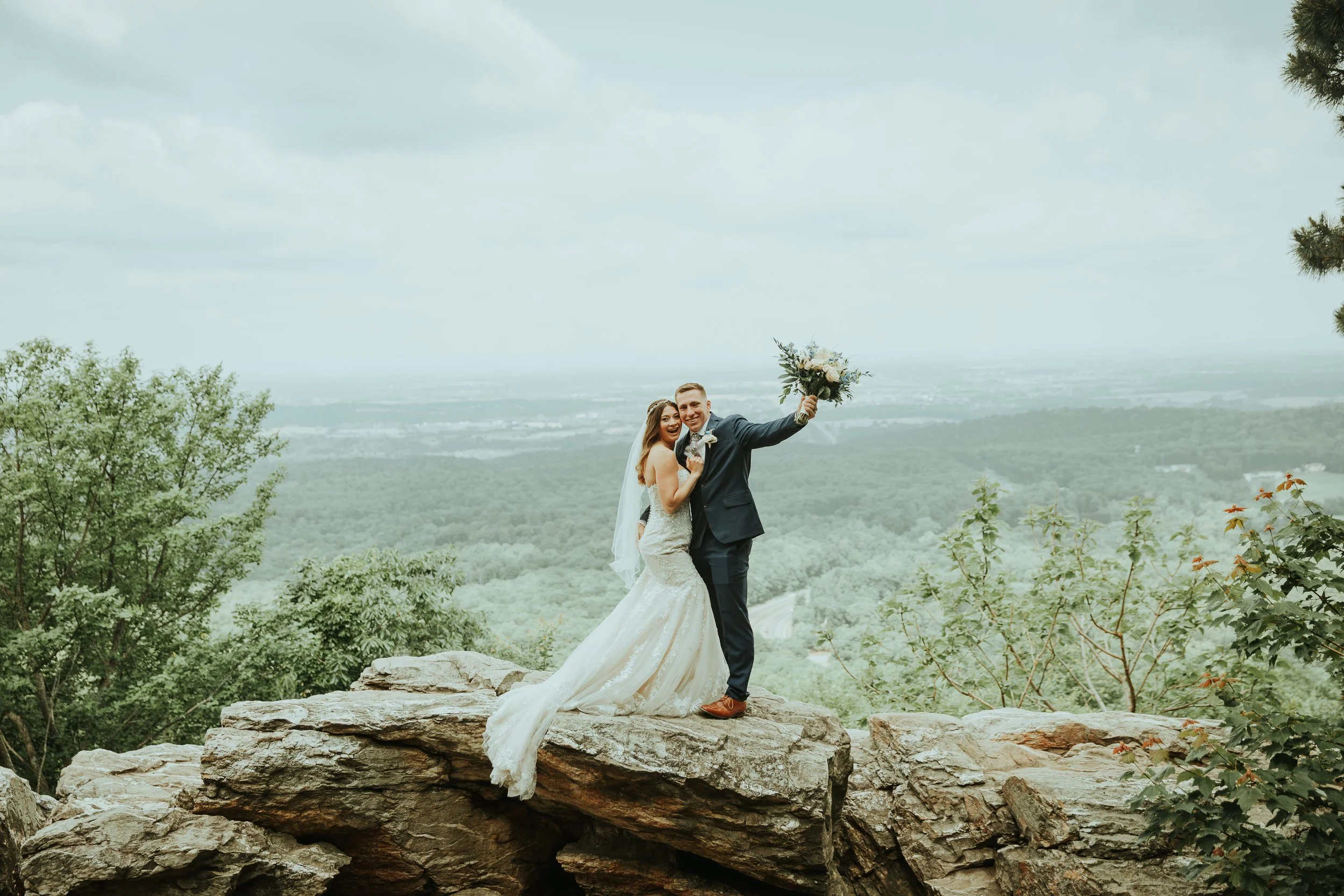 An elopement bride and groom standing on a rock formation outdoors with mountainous landscape in the background, celebrating their wedding day. The bride is wearing a white wedding gown and the groom is in a dark suit holding a bouquet of flowers.